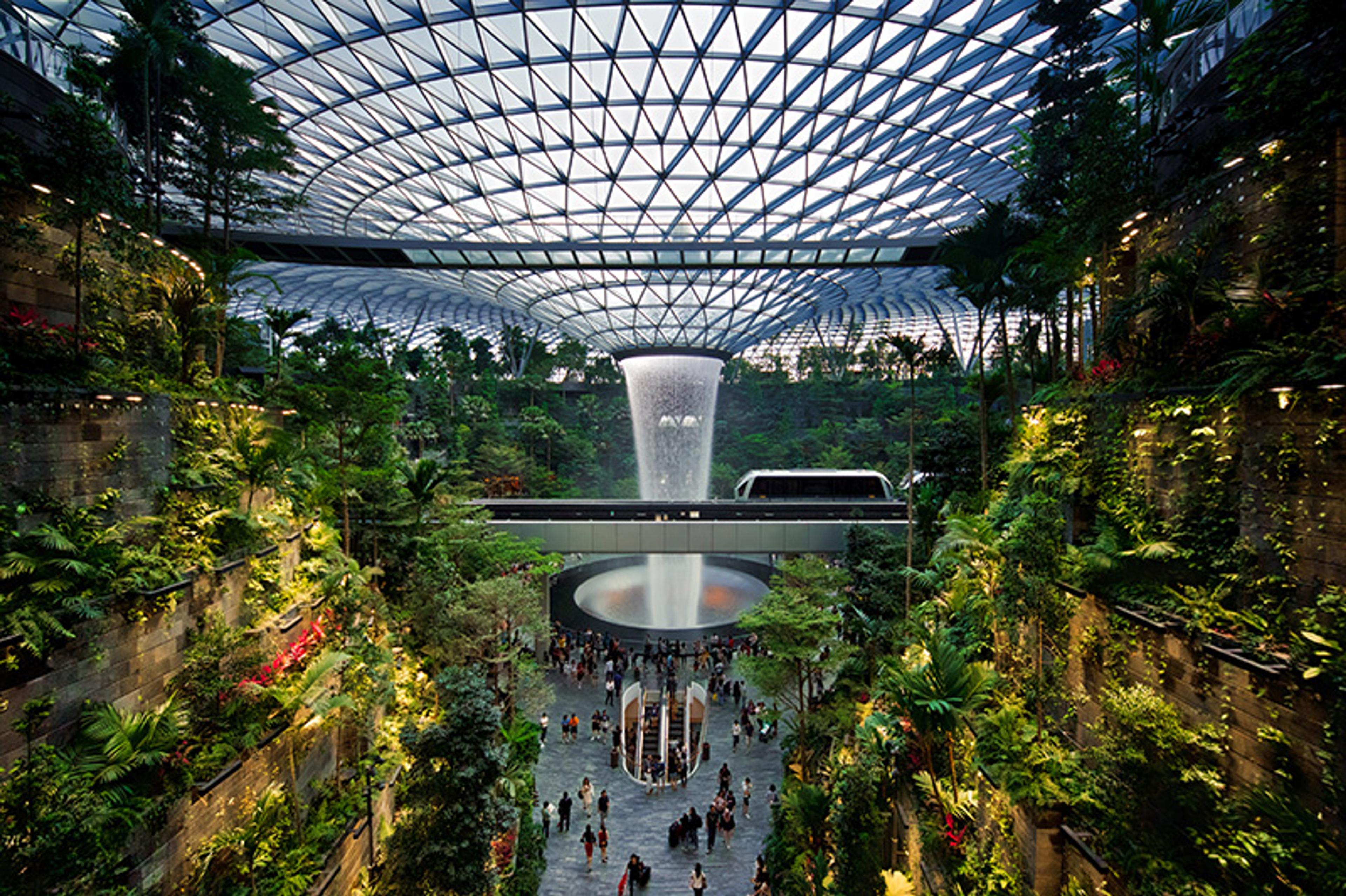 An indoor waterfall in a large glass dome with lush greenery and people walking below in a modern building.