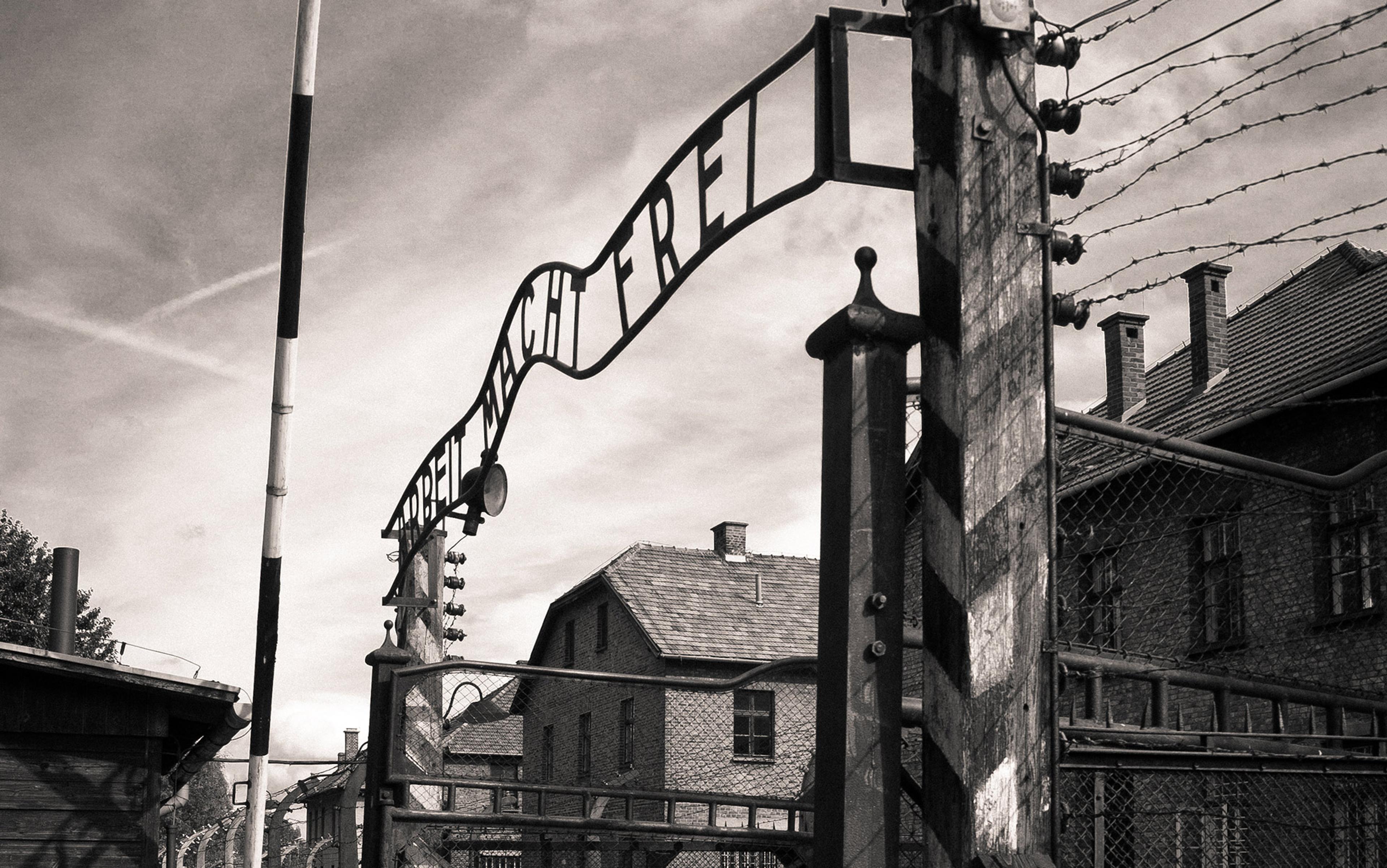 Black-and-white photo of Auschwitz entrance gate with “Arbeit macht frei” sign, barbed wire fences, and adjacent brick buildings.