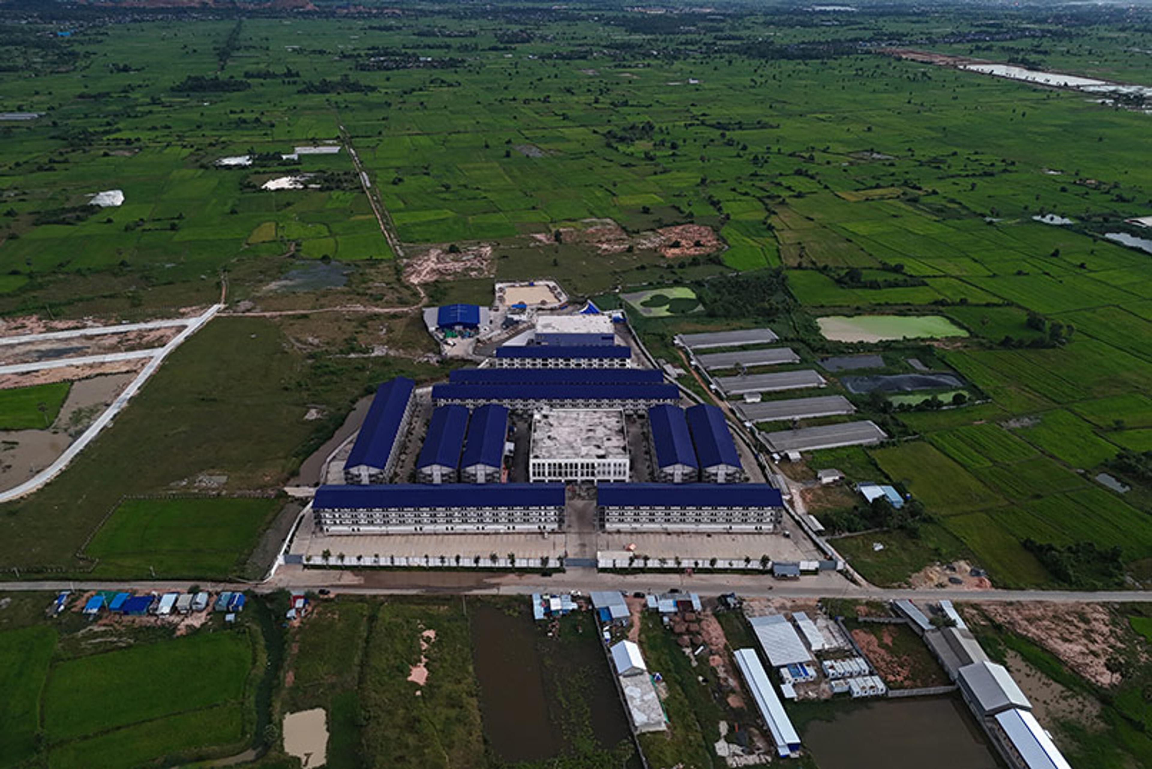 Aerial photo of a large industrial complex with blue roofs surrounded by green fields and scattered buildings.