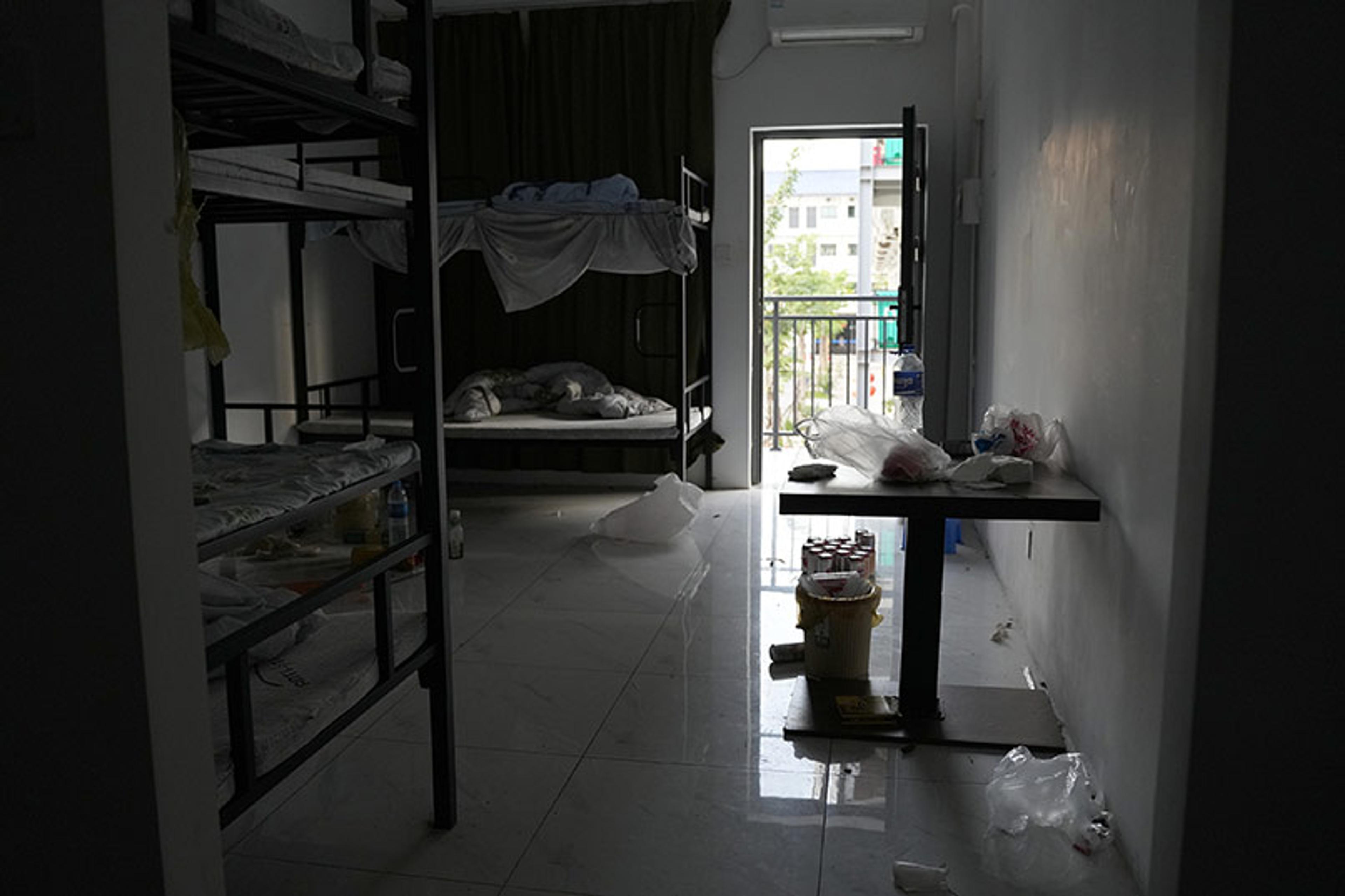 A messy dorm room with two sets of bunk beds and an open door leading to a balcony. Trash and clutter are on the floor and table.