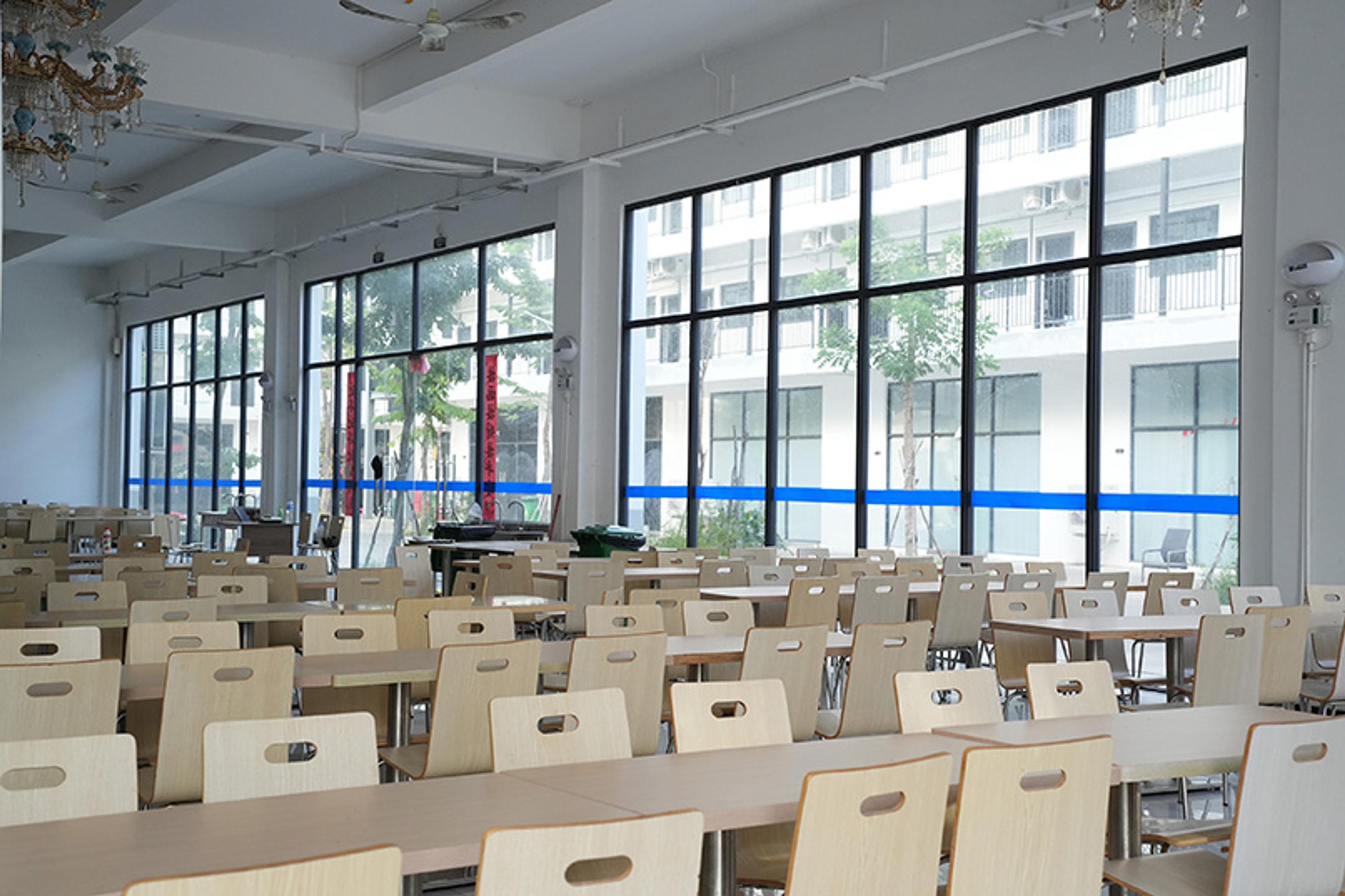 An empty canteen with wooden tables and chairs and large windows showing a neighbouring building.