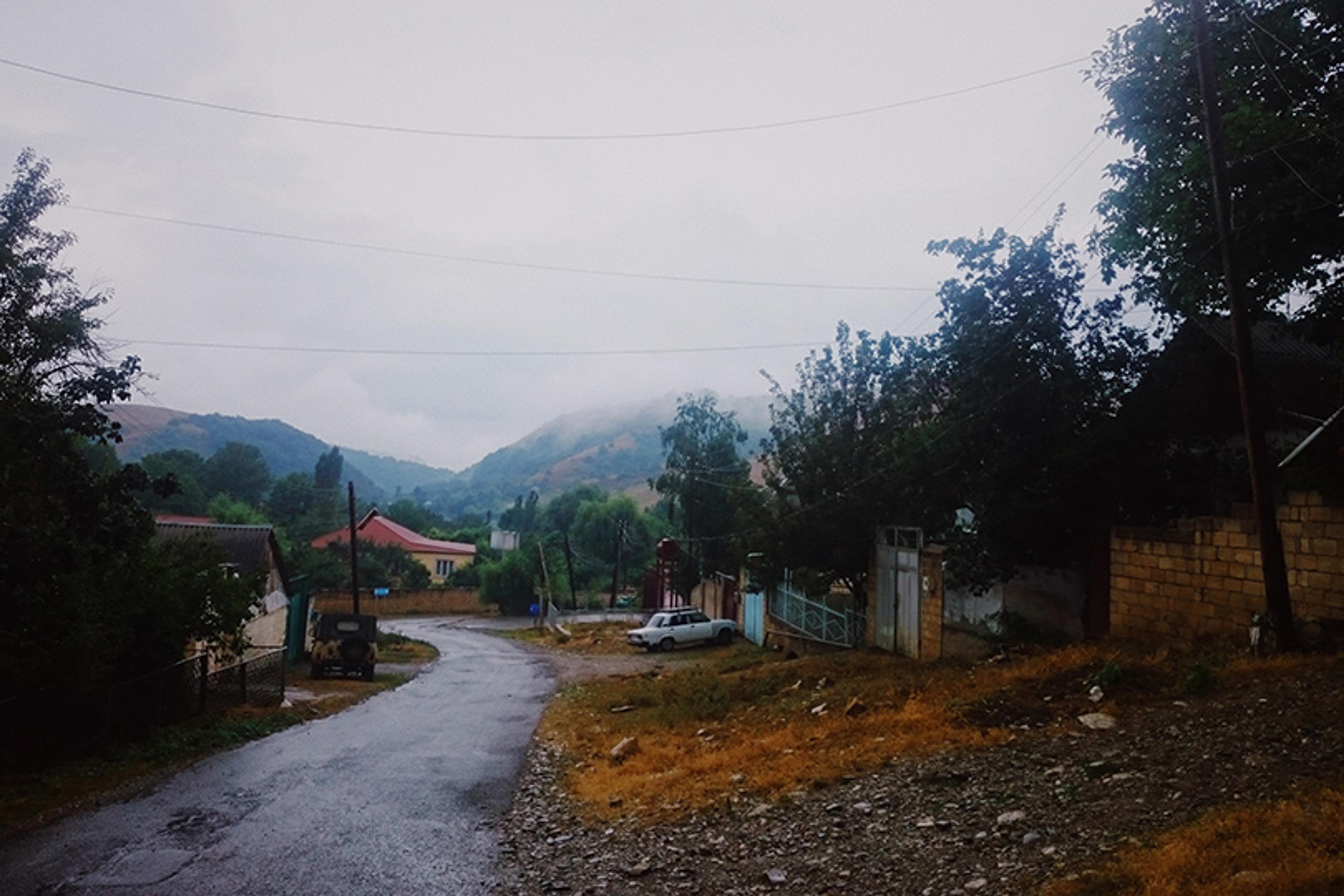 Photo of a rural road on a rainy day with houses, trees and misty hills in the background.