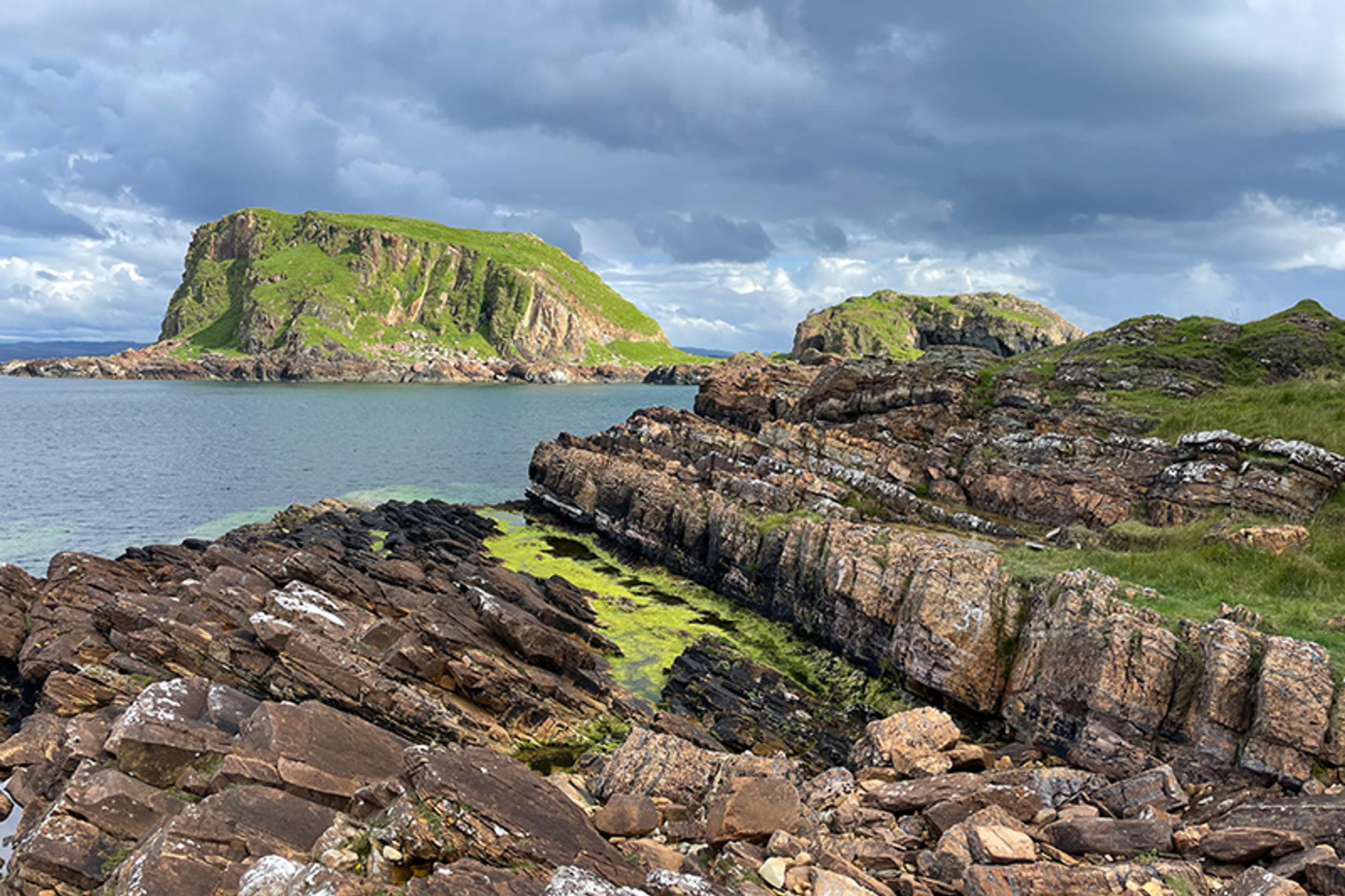 Photo of a rocky coastline with green cliffs and an overcast sky, featuring ocean views and rugged terrain.