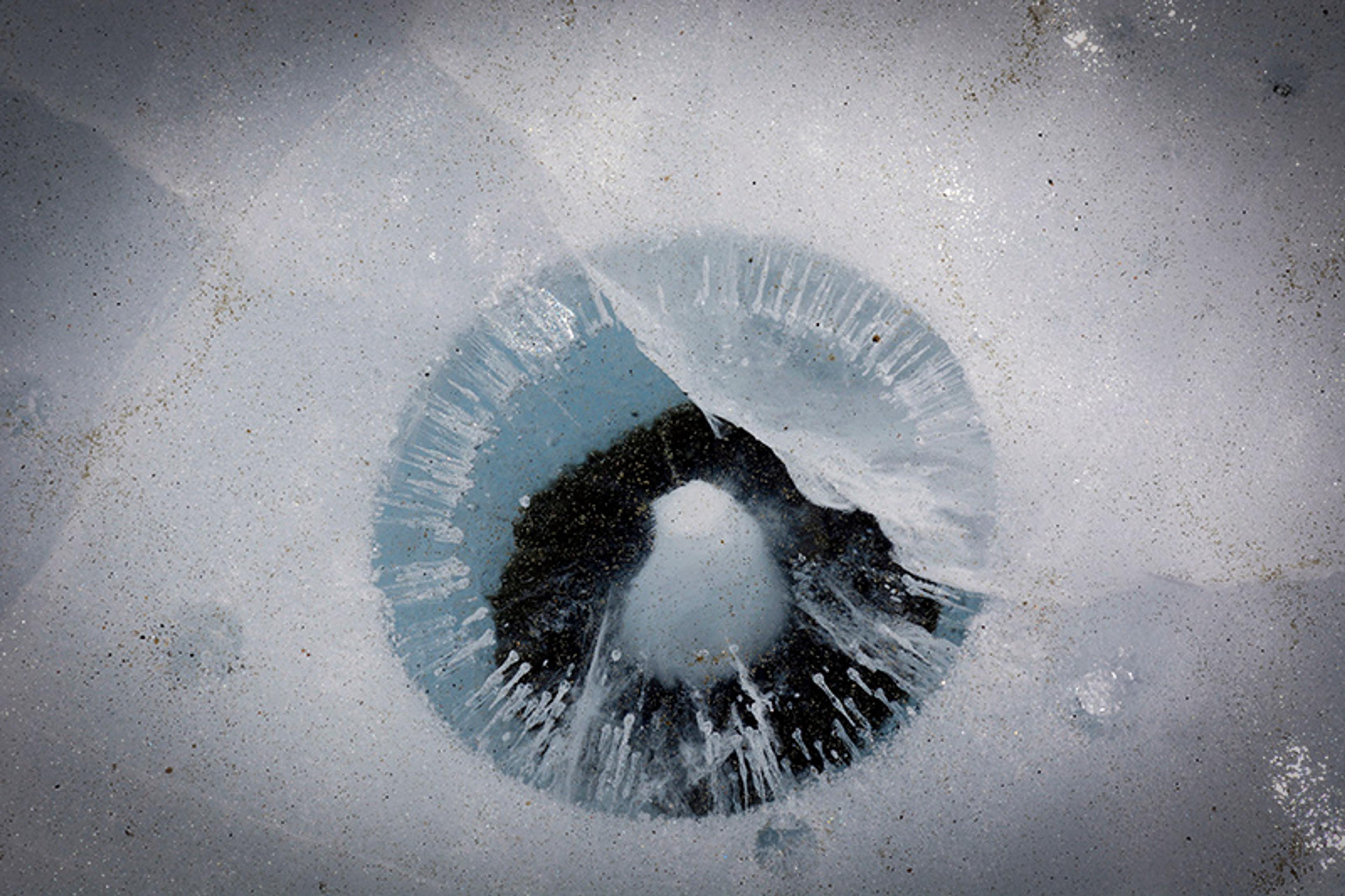 A circular ice formation resembling an eye on a frozen lake surface with cracks and bubbles visible.