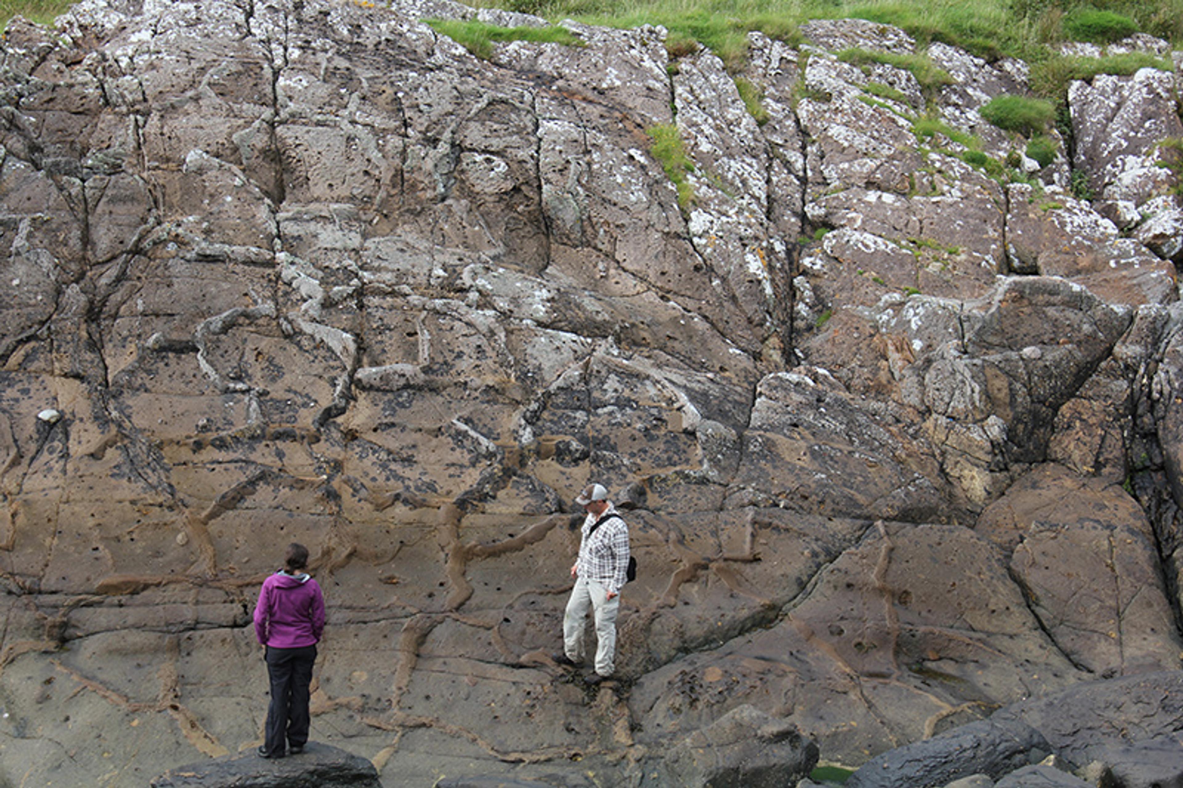 Two people standing on large rugged rocks with visible cracks and patches of greenery above.