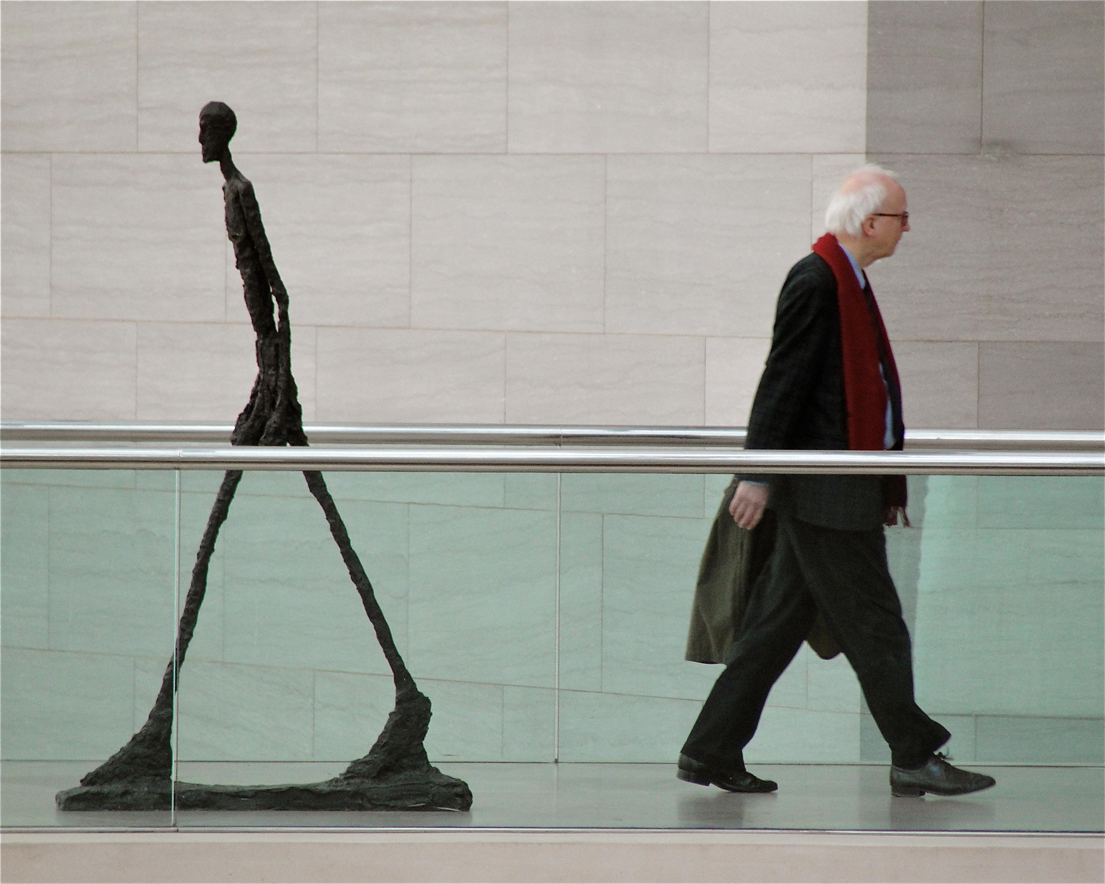 A thin sculpture walking in one direction and an elderly man with a red scarf walking in the other direction indoors.