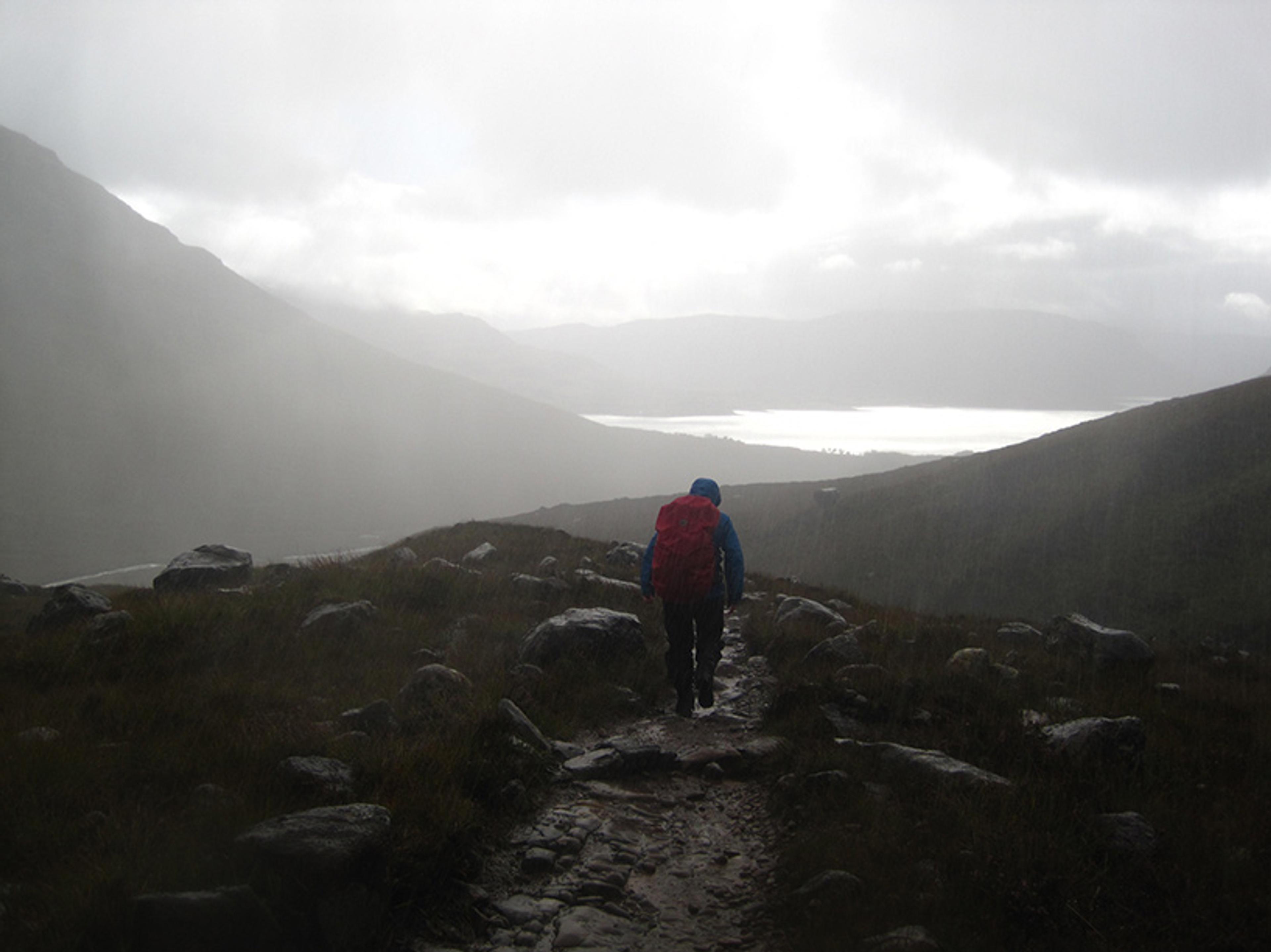 A person hiking on a rocky path among misty mountains with a red backpack and overcast skies.