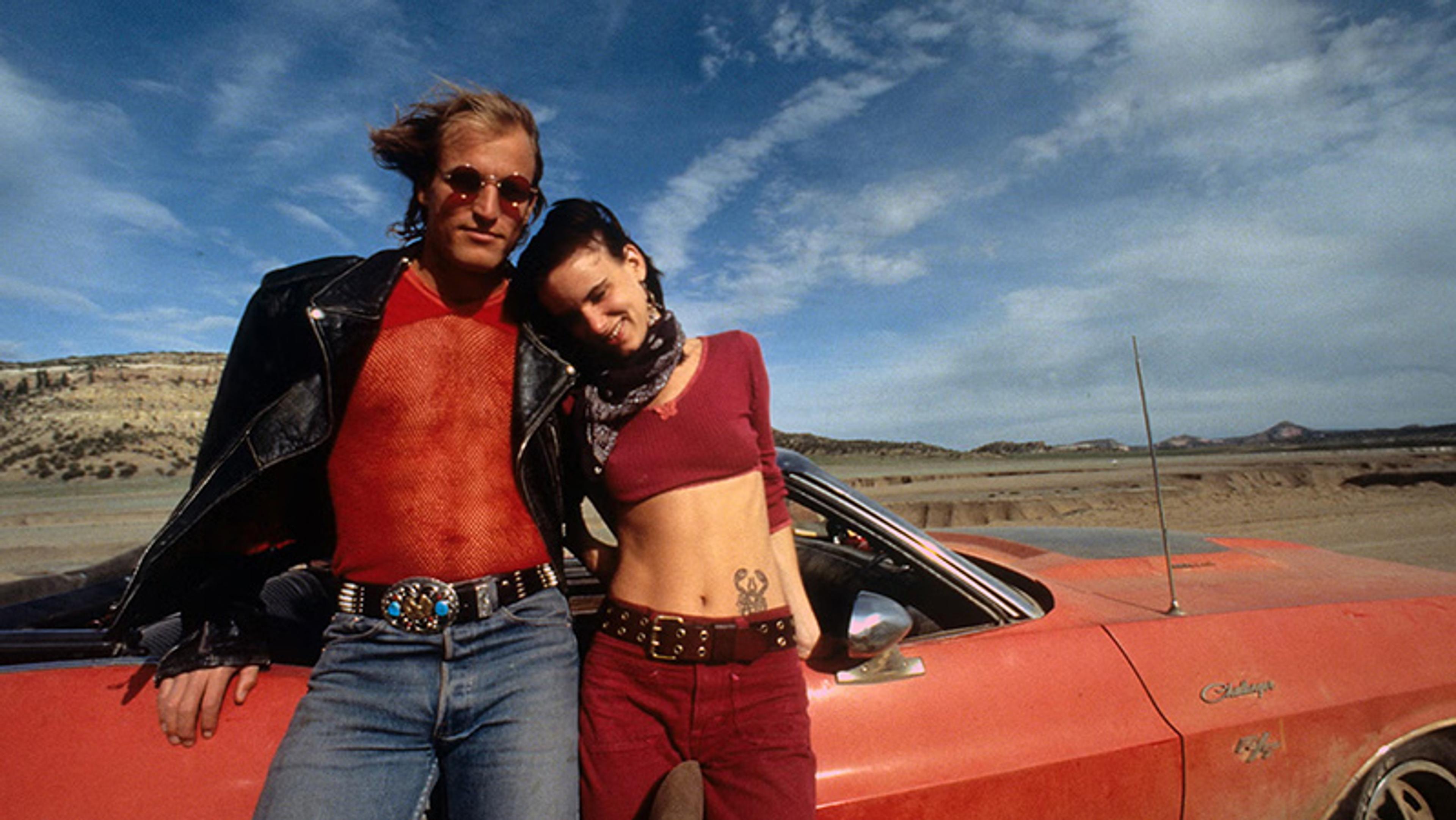 A man and woman leaning on a red car in a desert landscape under a blue sky.