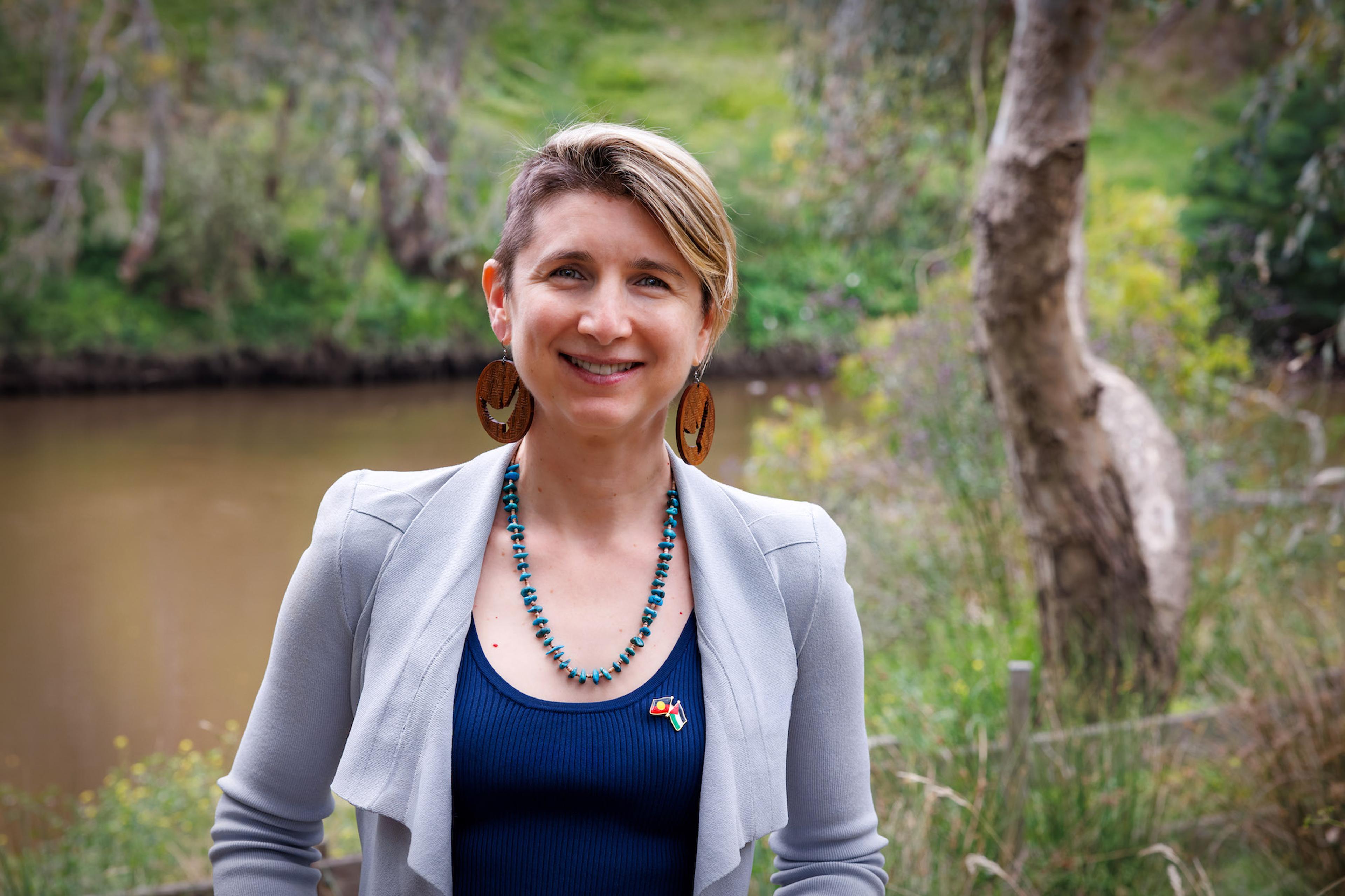 Photo of a smiling person with short hair standing outdoors near a river and trees.