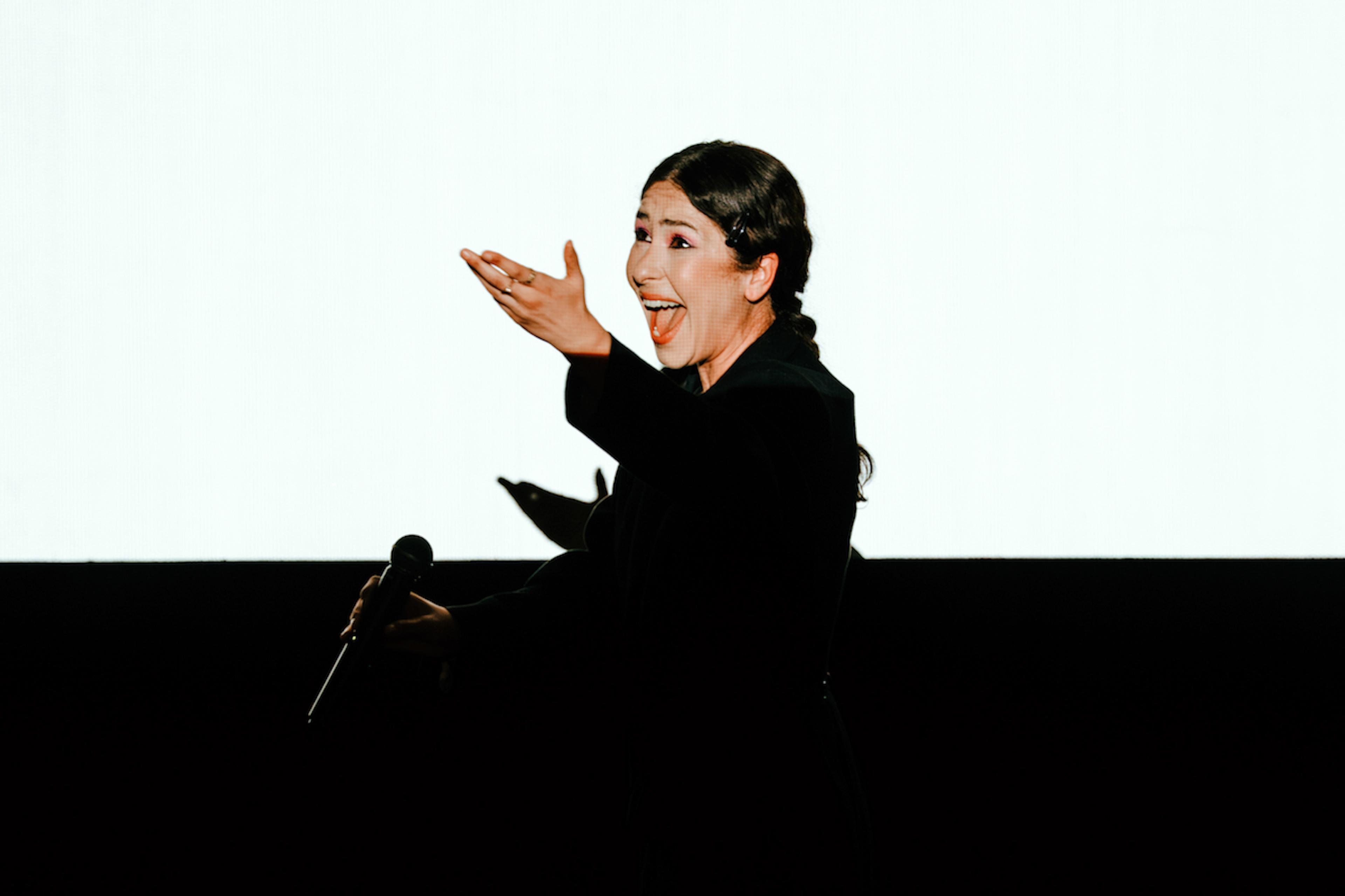 Photo of a person in a black outfit holding a microphone, singing expressively against a bright background.