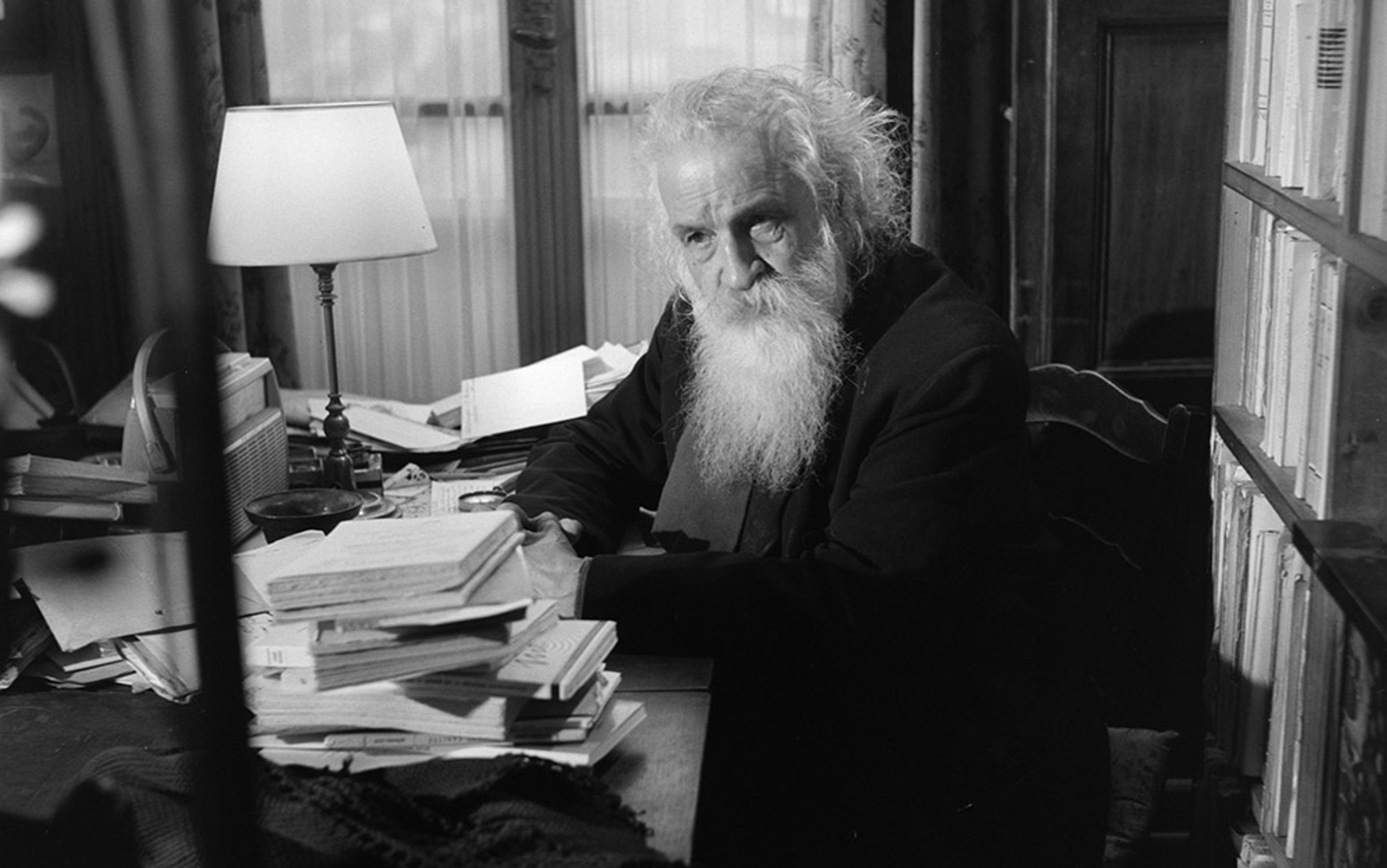 Vintage photo of an elderly man with a long white beard sitting at a cluttered desk in a book-filled study.