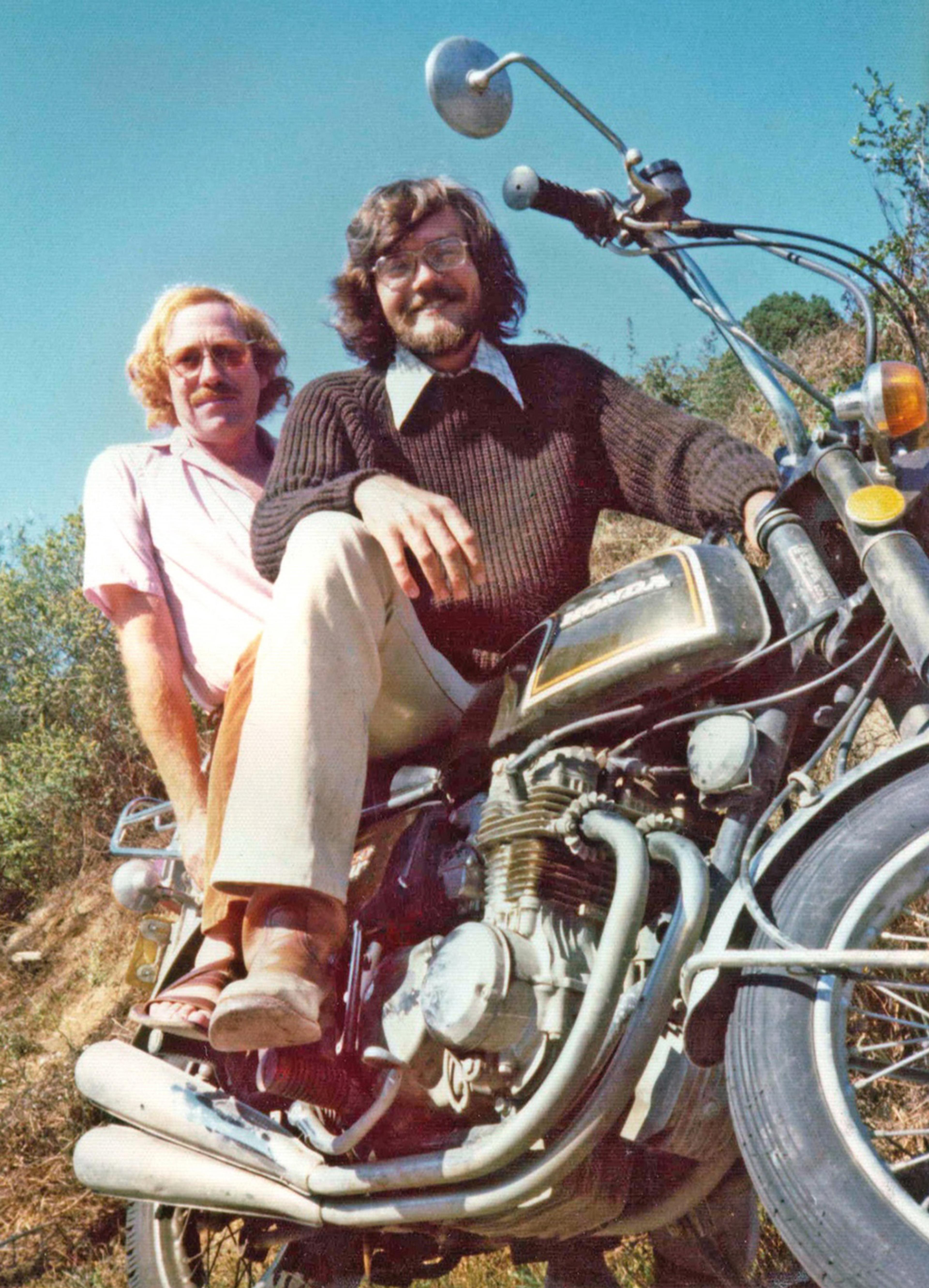 Two men sitting on a vintage motorcycle in a sunny outdoor setting with clear blue sky and greenery in the background.