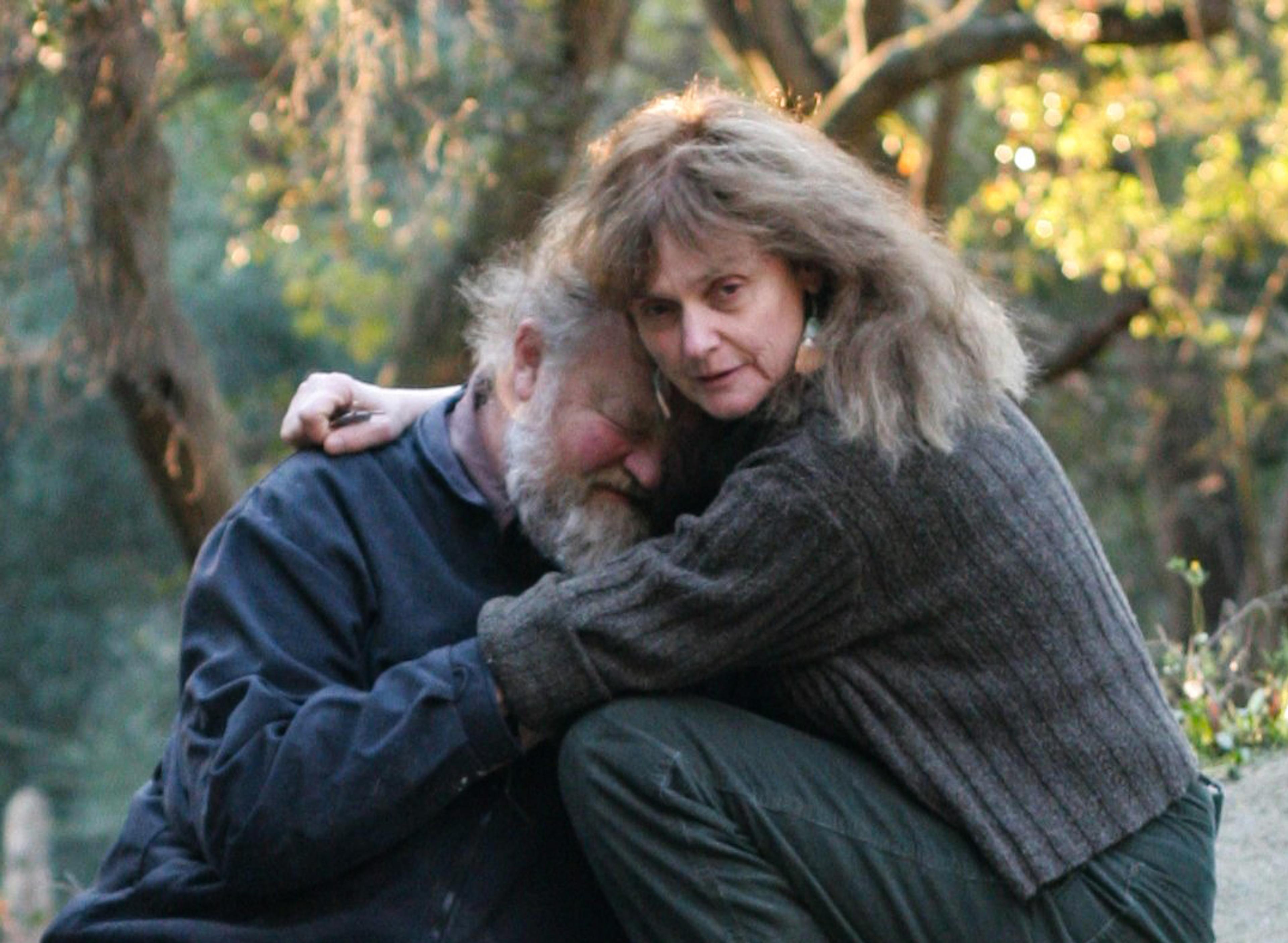 A woman embracing a bearded man in a forest. Both are seated on the ground with trees softly lit by sunlight.
