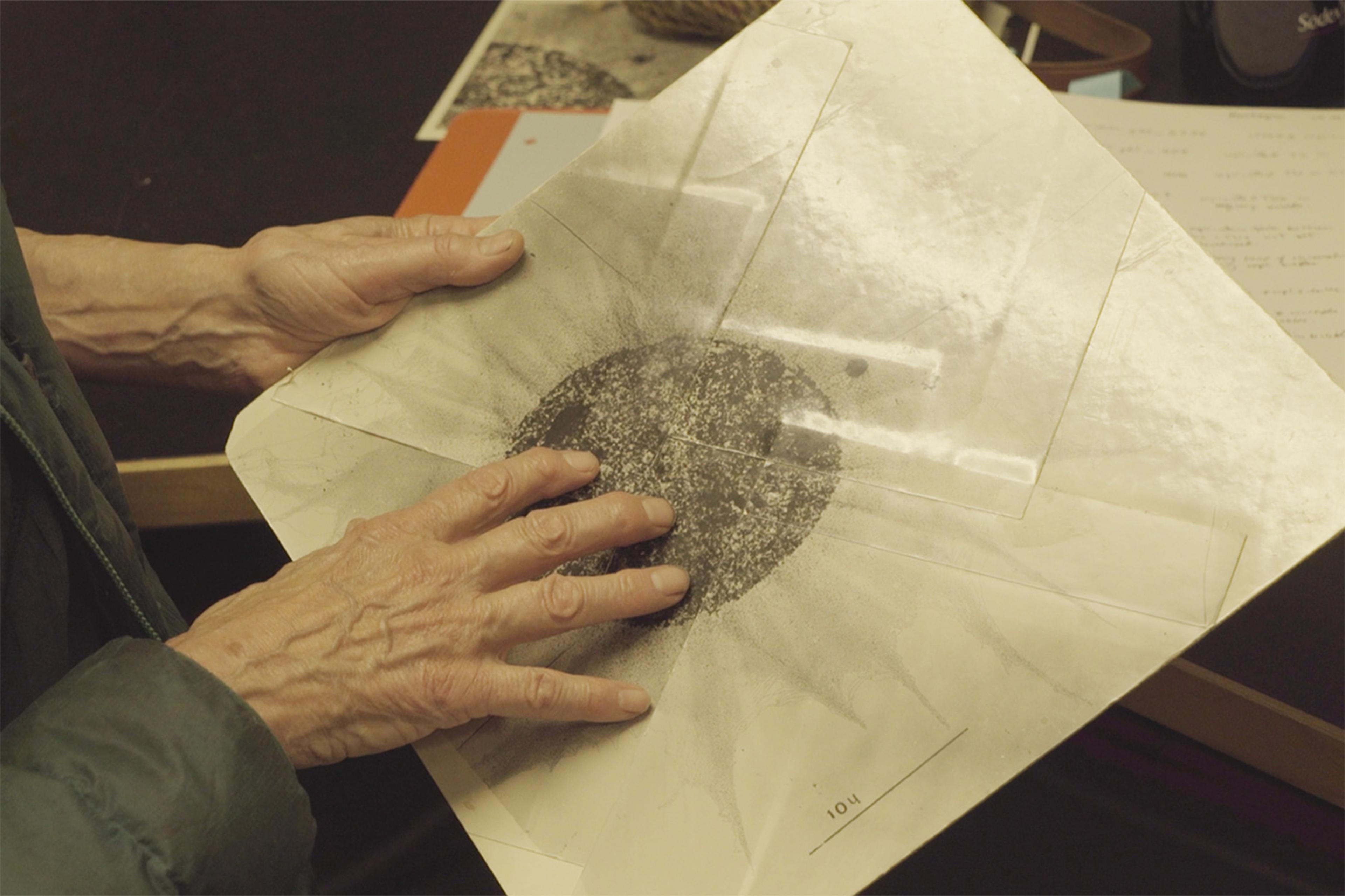 A person’s hand touching a vintage document with a central black pattern on a table with papers and items.