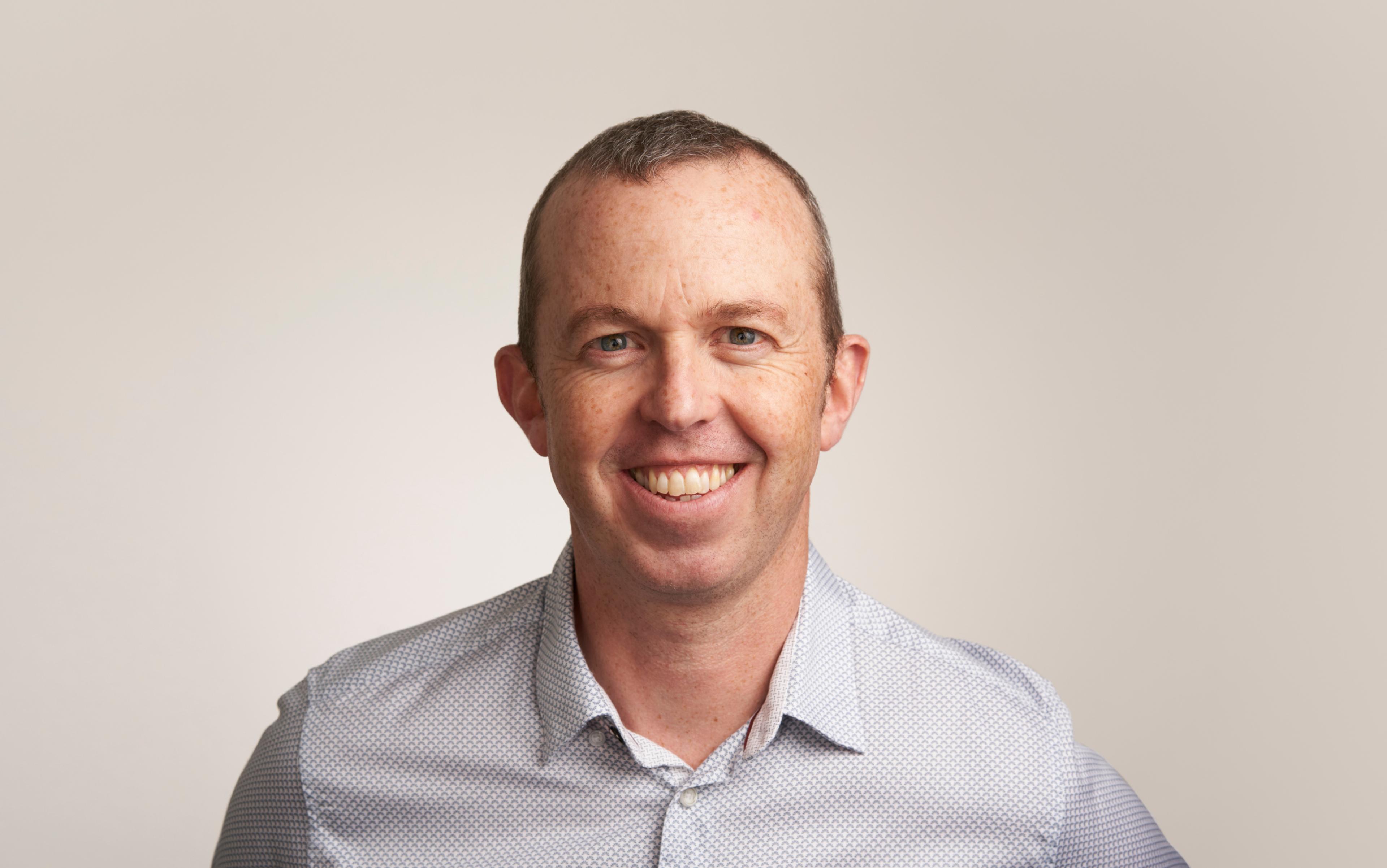 Photo of a smiling man wearing a light blue patterned shirt against a neutral background.