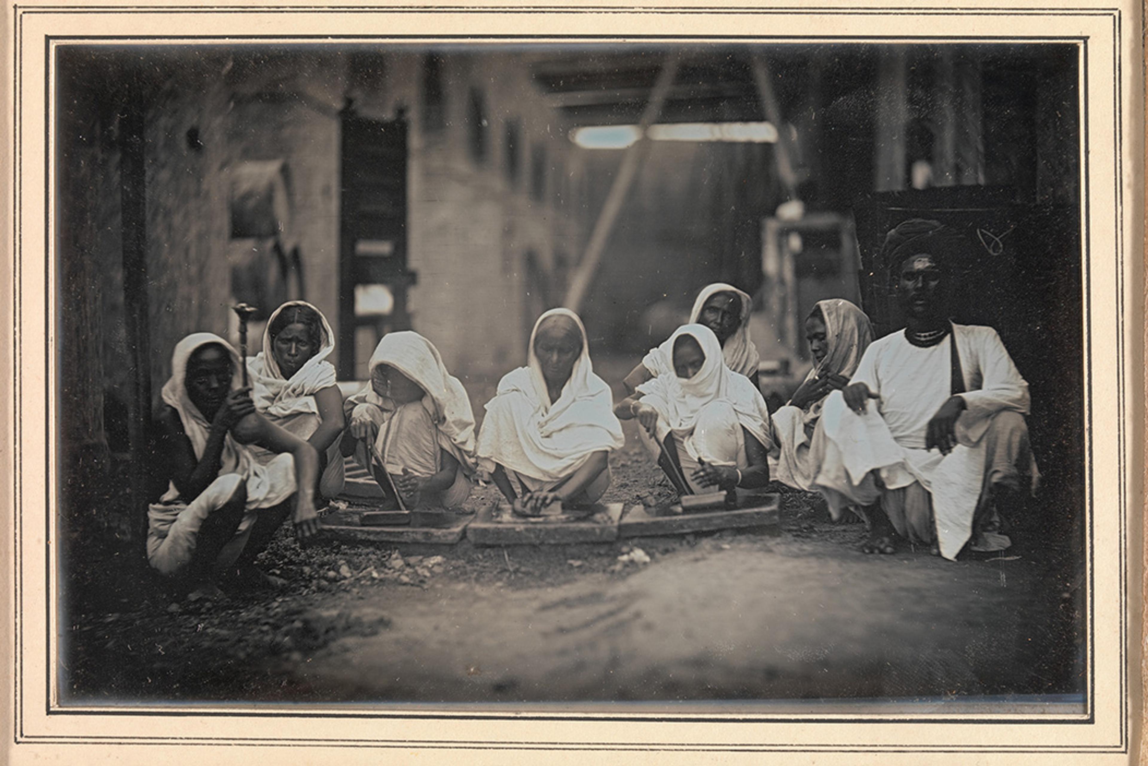 Vintage photo of people in traditional attire seated in a row, working with tools in an industrial setting.