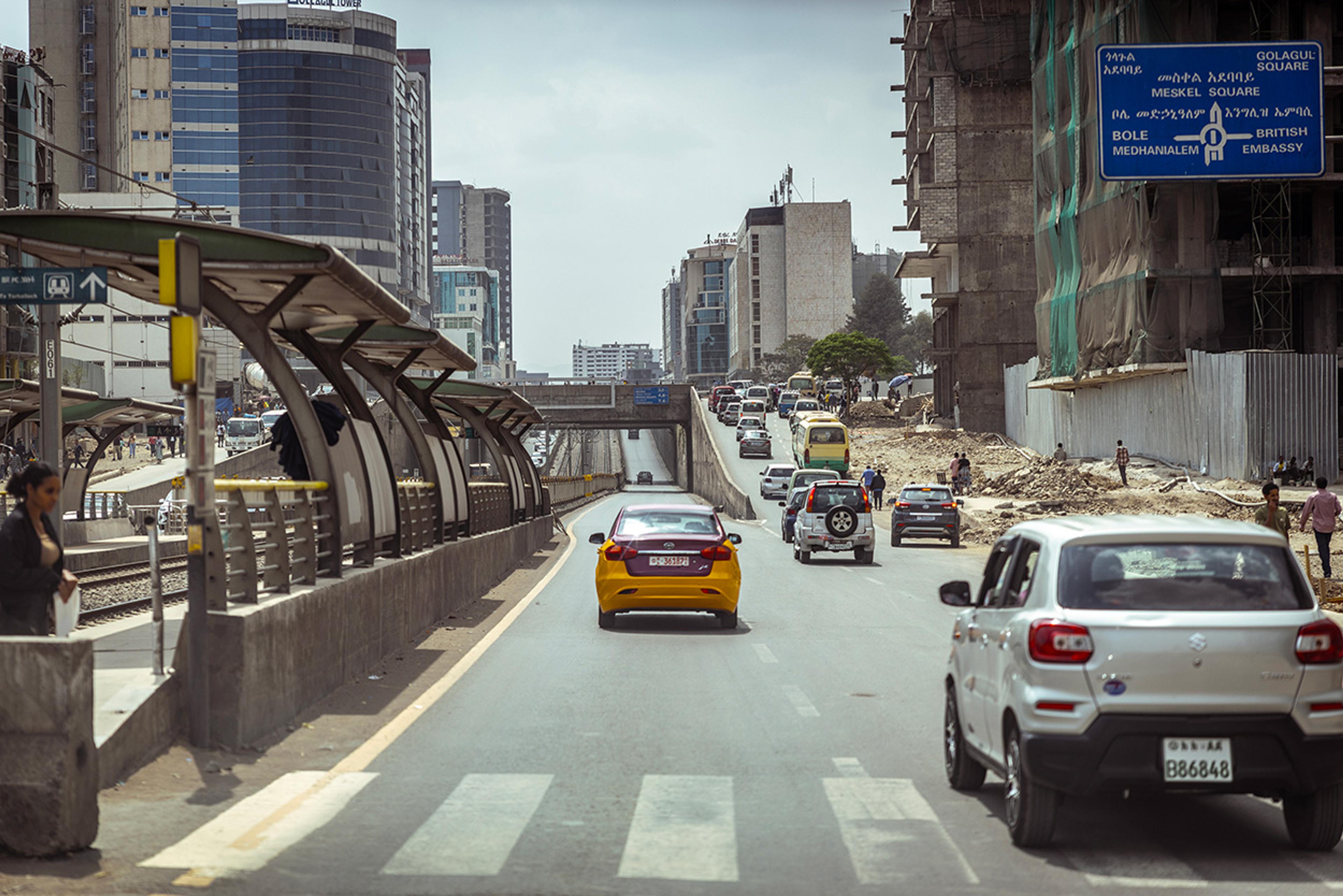 An urban street with vehicles, a pedestrian standing on a platform, high-rise buildings and a blue road sign.