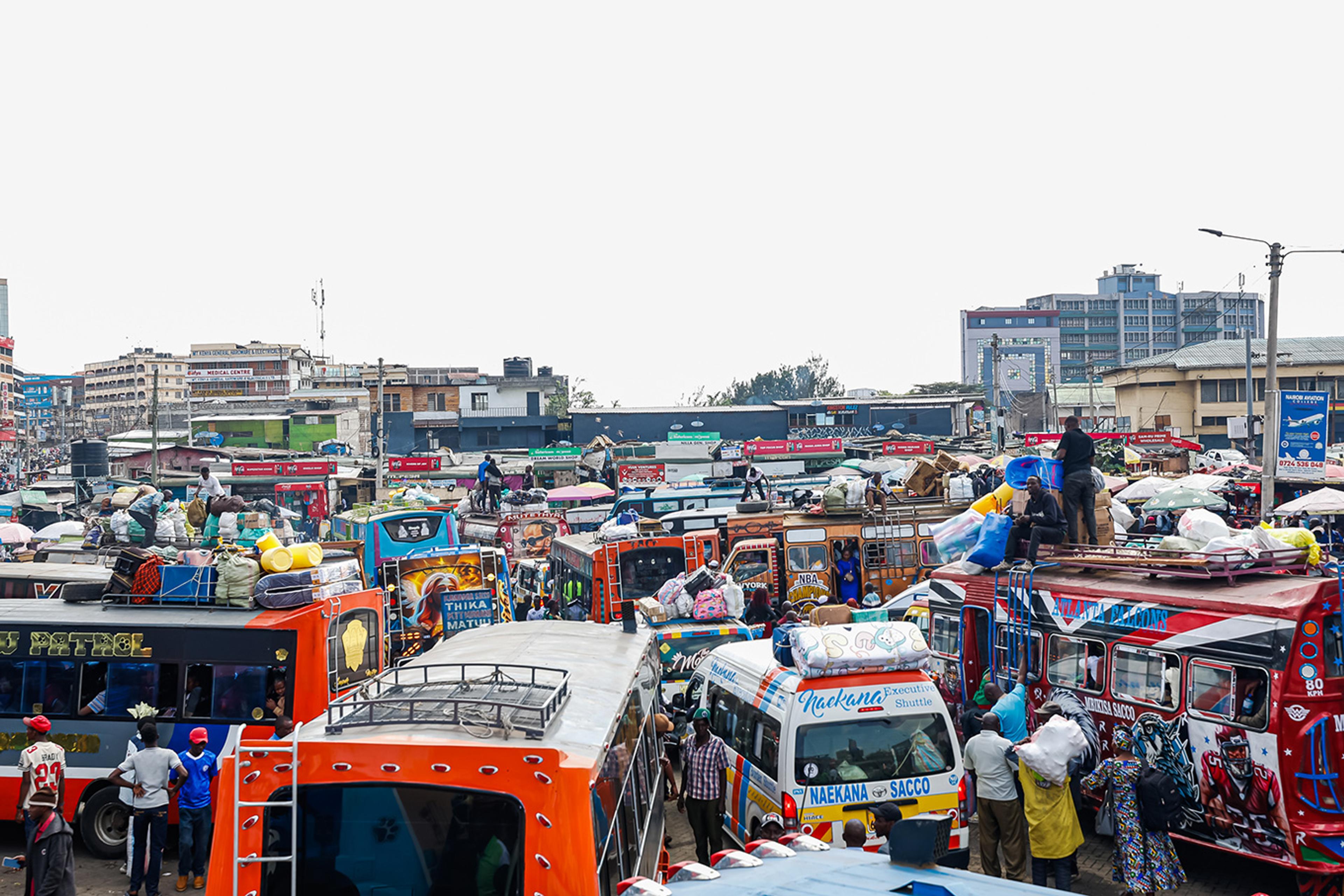 A busy urban bus terminal with colourful buses, people loading luggage and city buildings in the background.