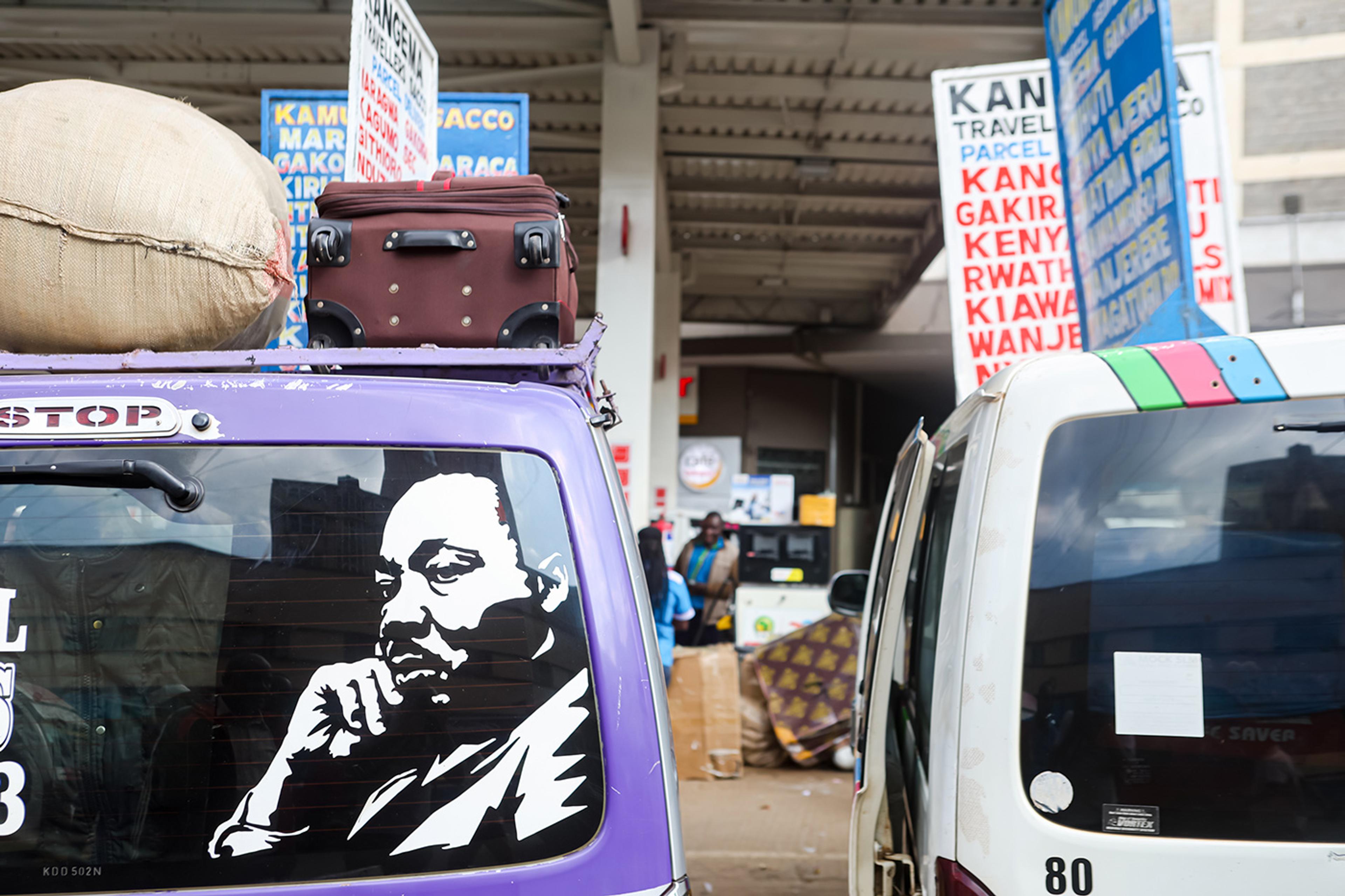 A purple van with a portrait decal, luggage on top, parked at a bus station with signs and people in the background.