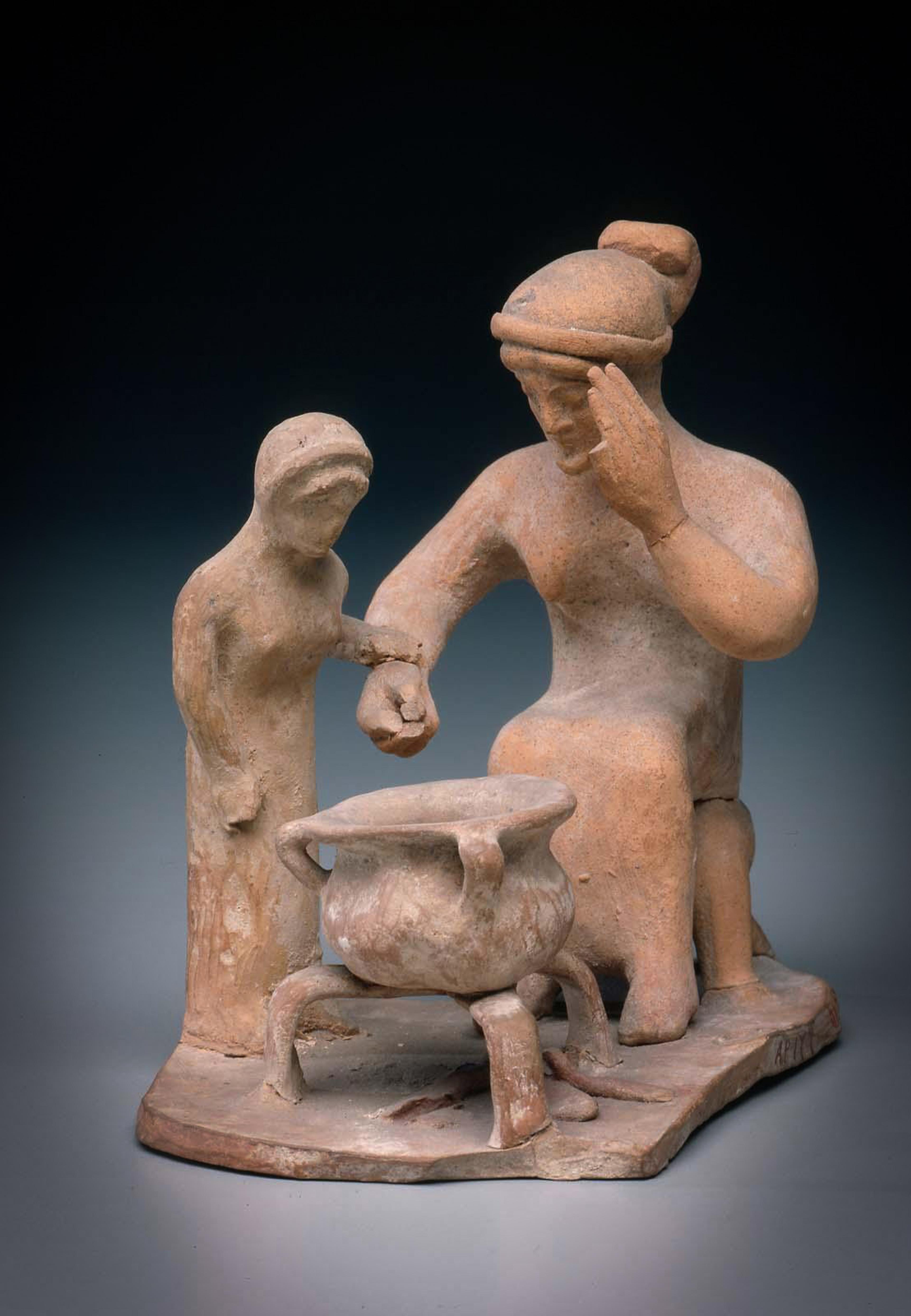 Ancient clay sculpture of two figures around a pot on a fire, one seated, wearing a head wrap with a hand raised to face.