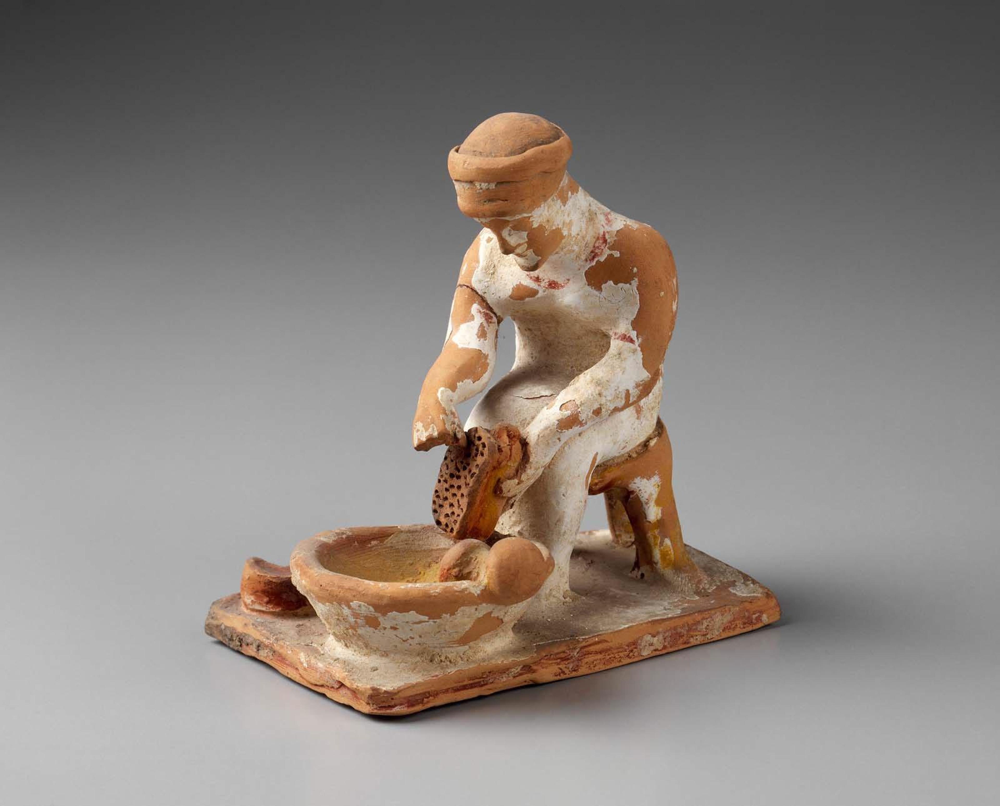 Ancient terracotta sculpture of a seated woman grating cheese into a basin, worn with age against a grey background.