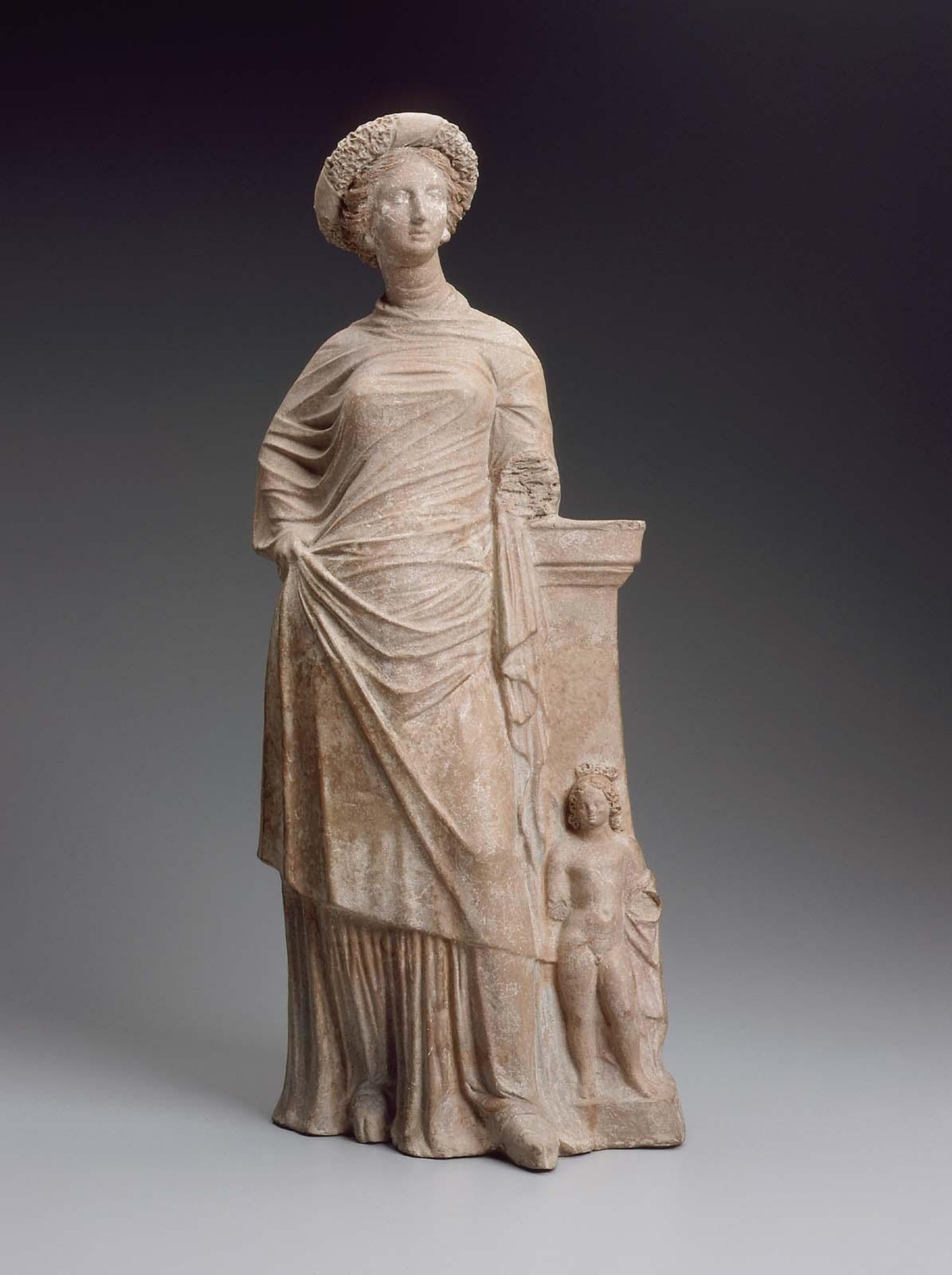 Ancient marble statue of a woman in draped clothing beside a small child figure on a grey background.