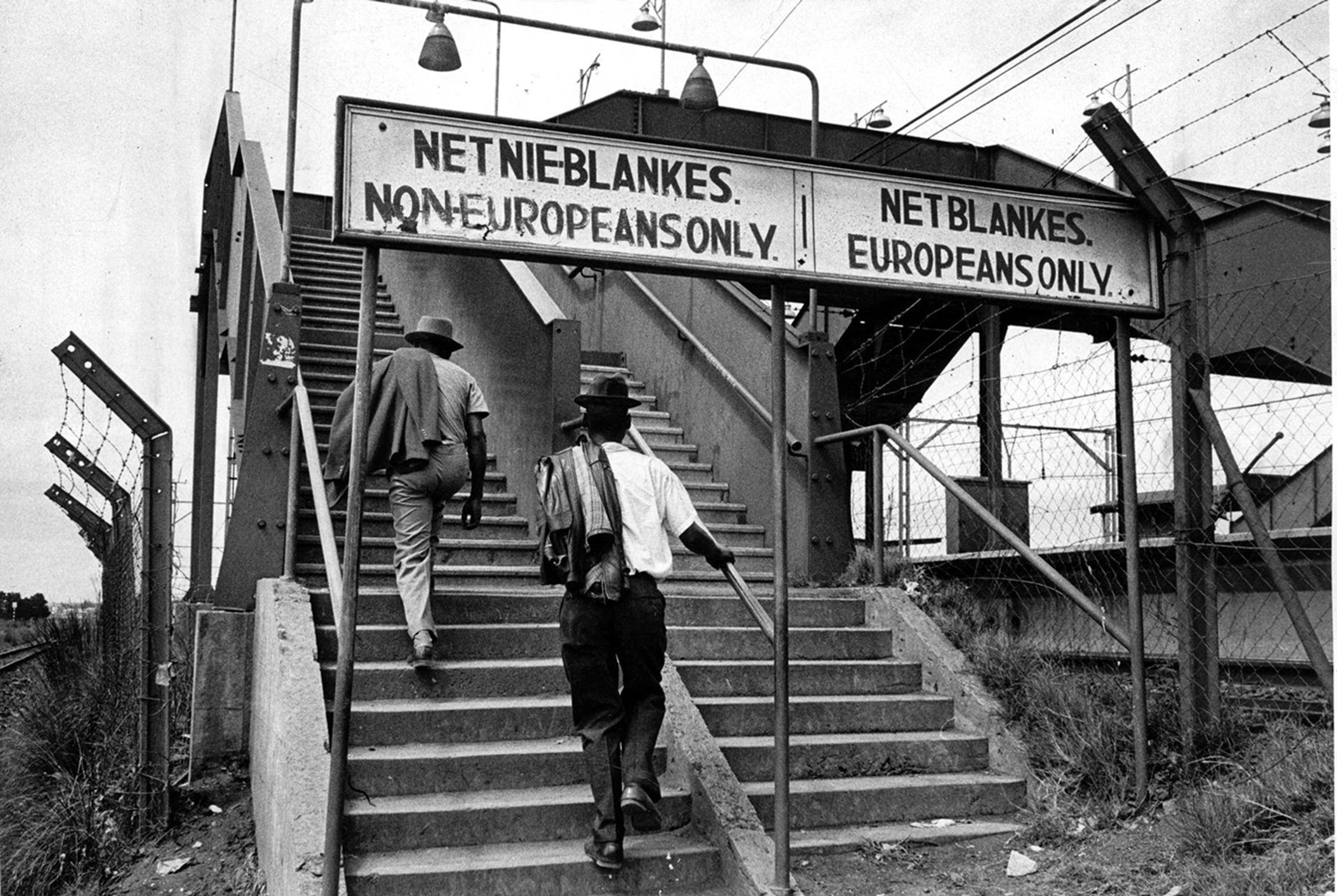 Black-and-white photo of two Black men ascending a segregated stairway beneath the sign 'Non-Europeans Only' at a train station in South Africa.