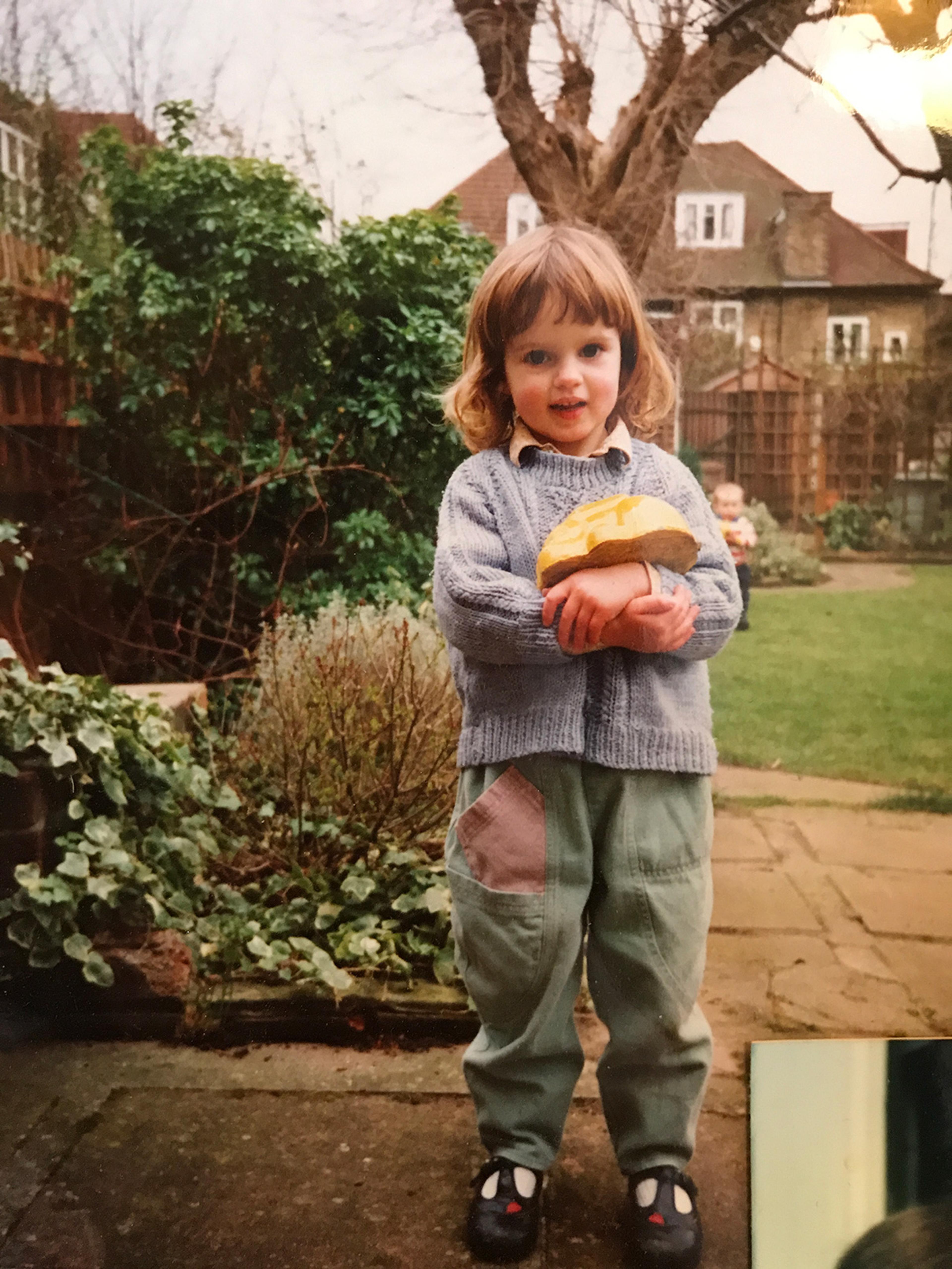 Photo of a girl in a garden wearing a jumper and patched trousers with a house and trees in the background.