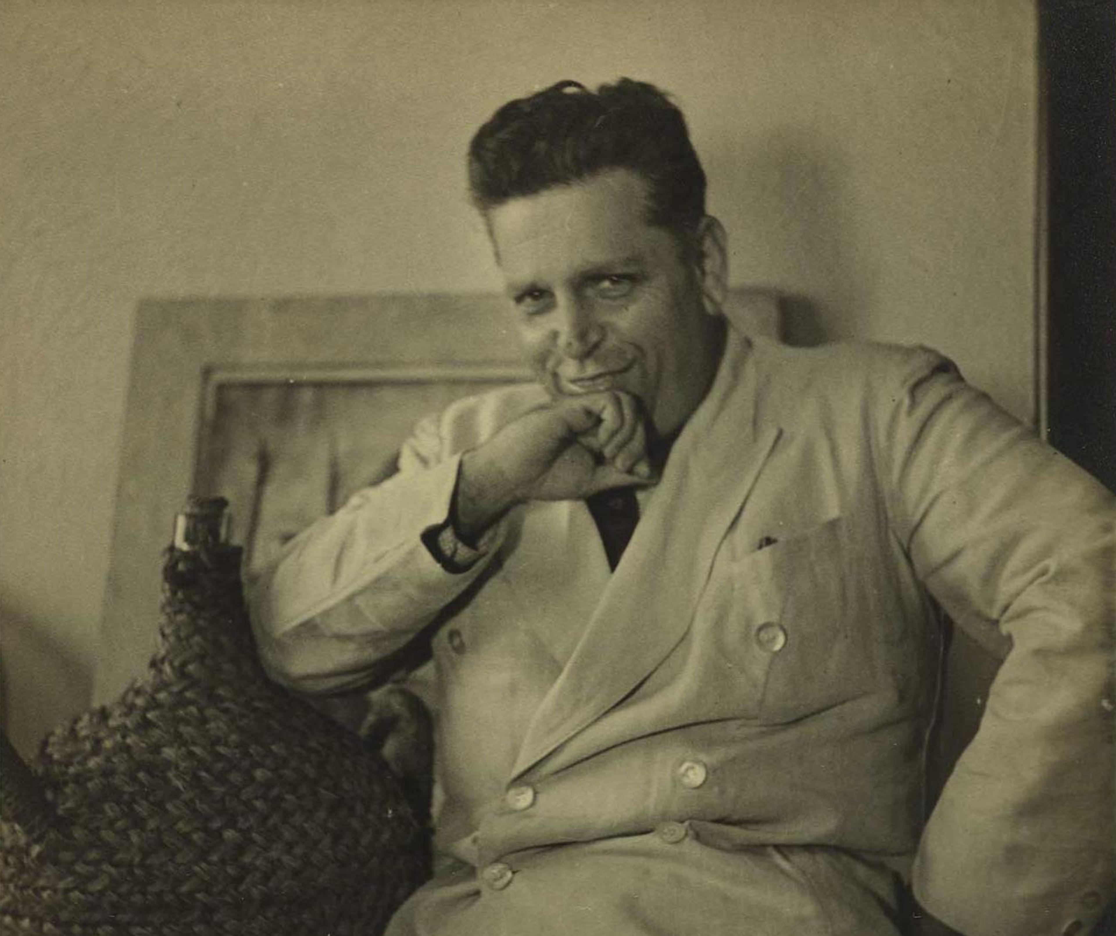 Black and white photo of a man in a suit sitting, hand on chin, with a woven basket beside him.