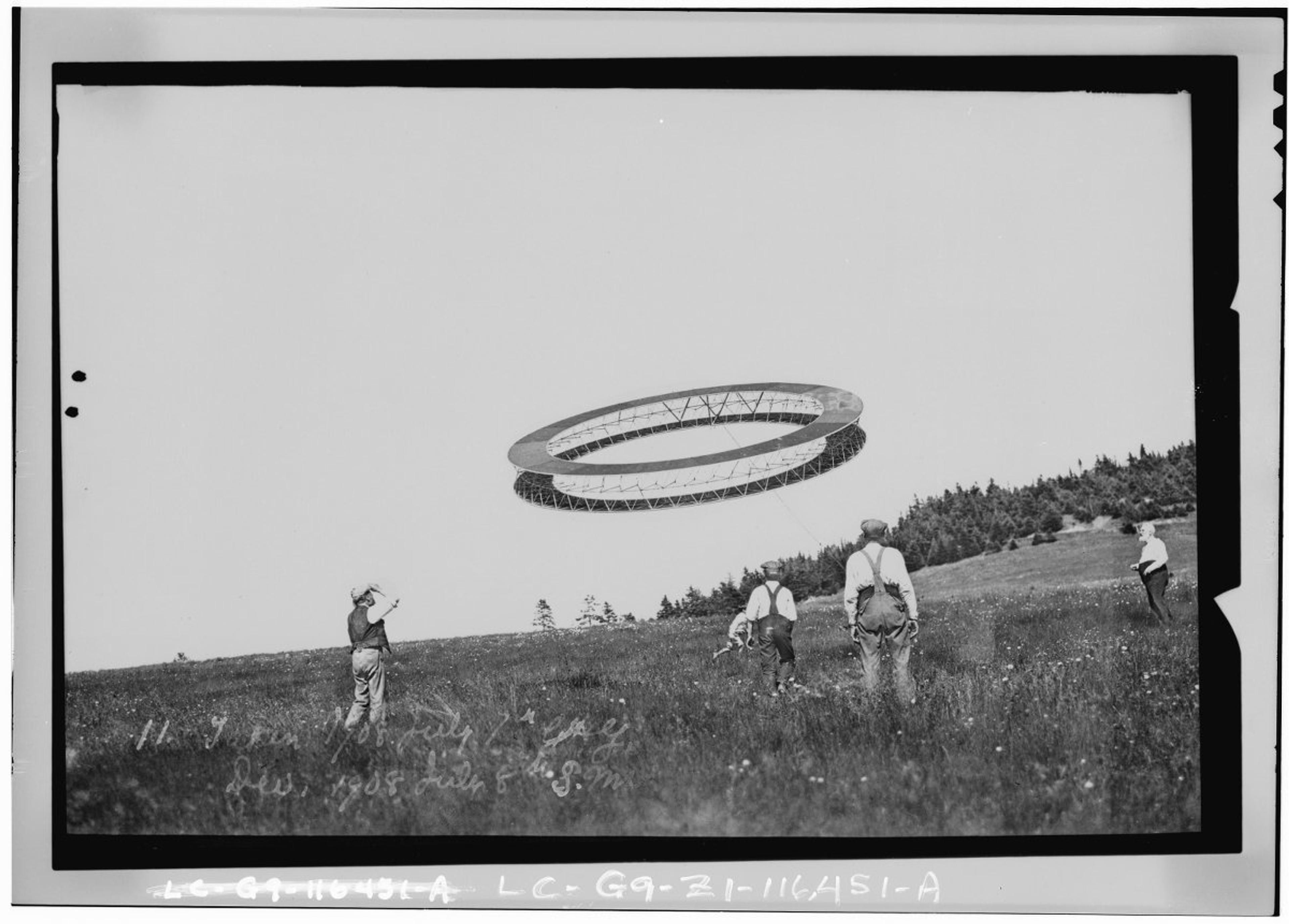 Black and white photo of a group of people observing a large, ring-shaped object flying in a field.