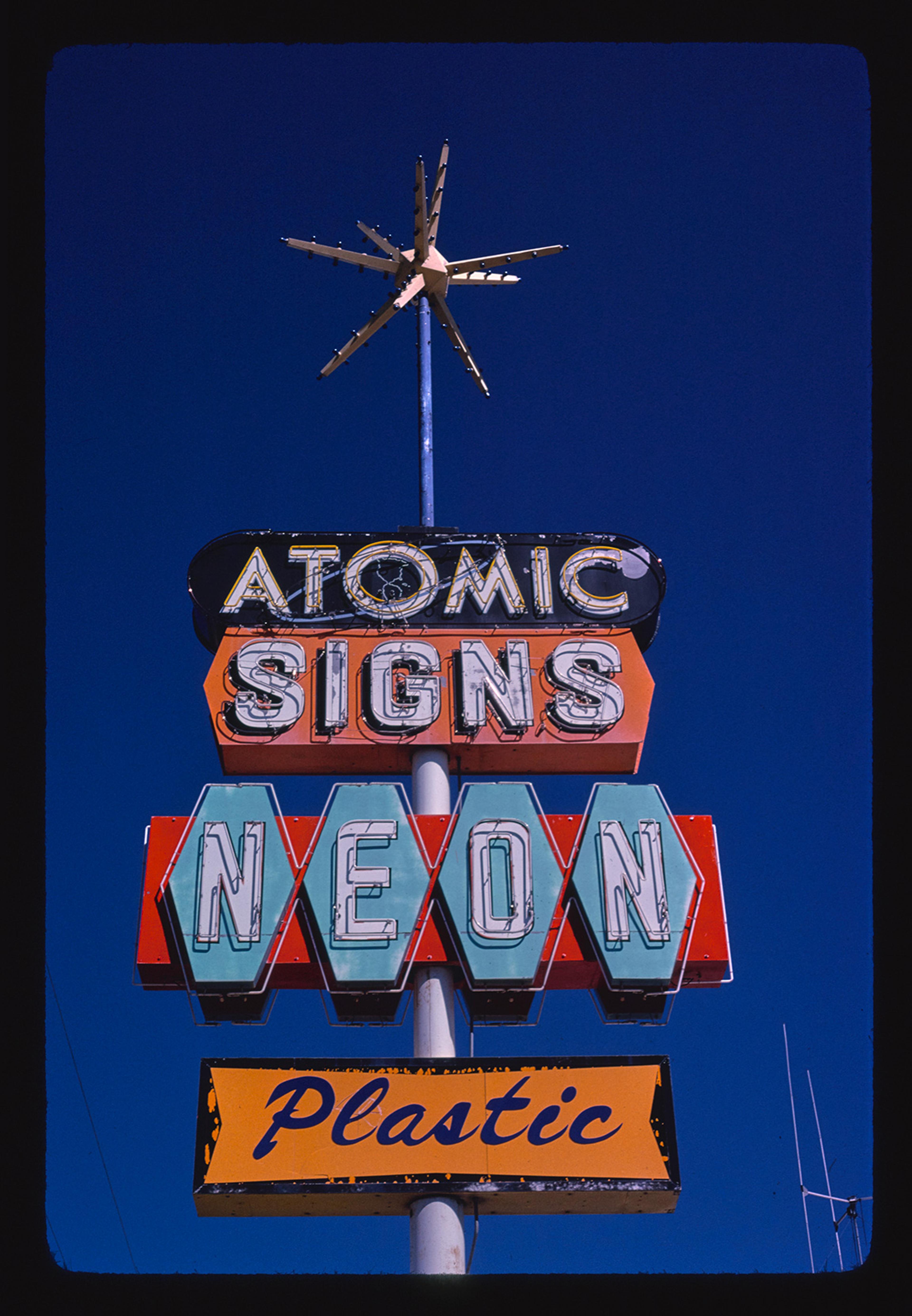 A vintage neon sign reading “Atomic Signs Neon Plastic” against a clear blue sky, with a starburst ornament on top.