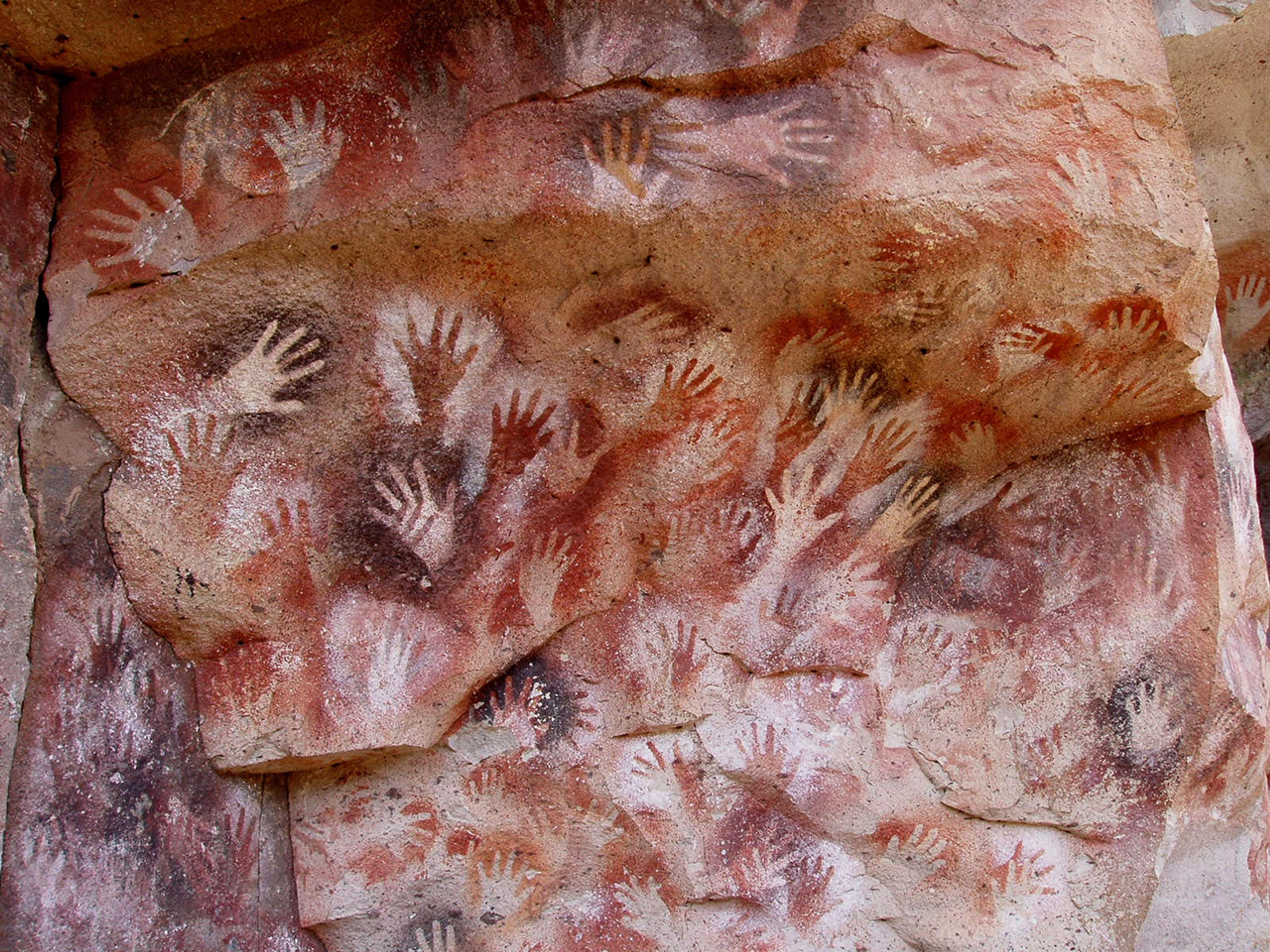 Painting of ancient handprints in various shades on a rocky surface, with a red and brown background.