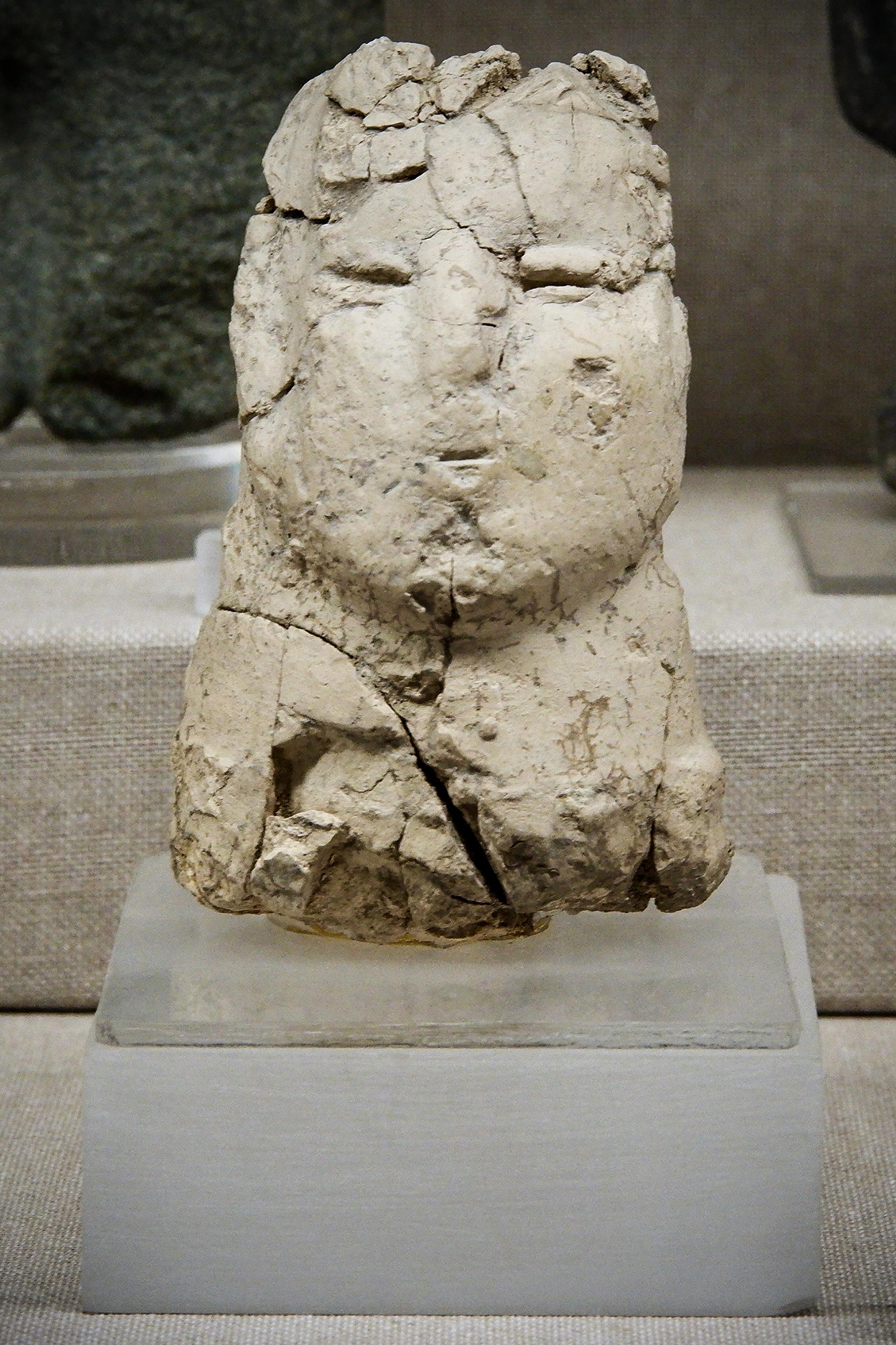 A weathered, ancient clay sculpture of a human face on a display pedestal in a museum setting.
