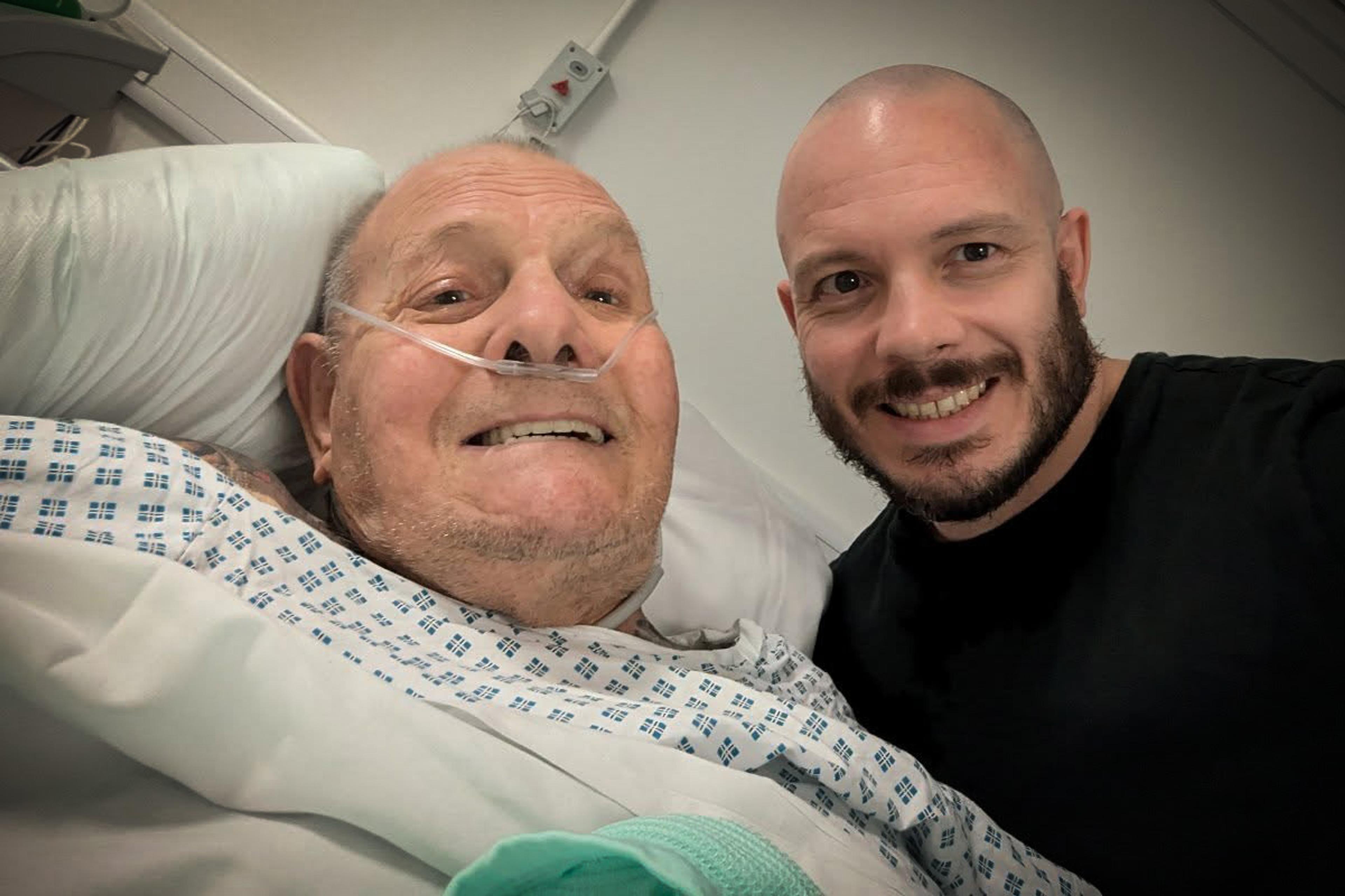 An elderly man in a hospital bed smiling with a younger man beside him, both appearing cheerful and relaxed.