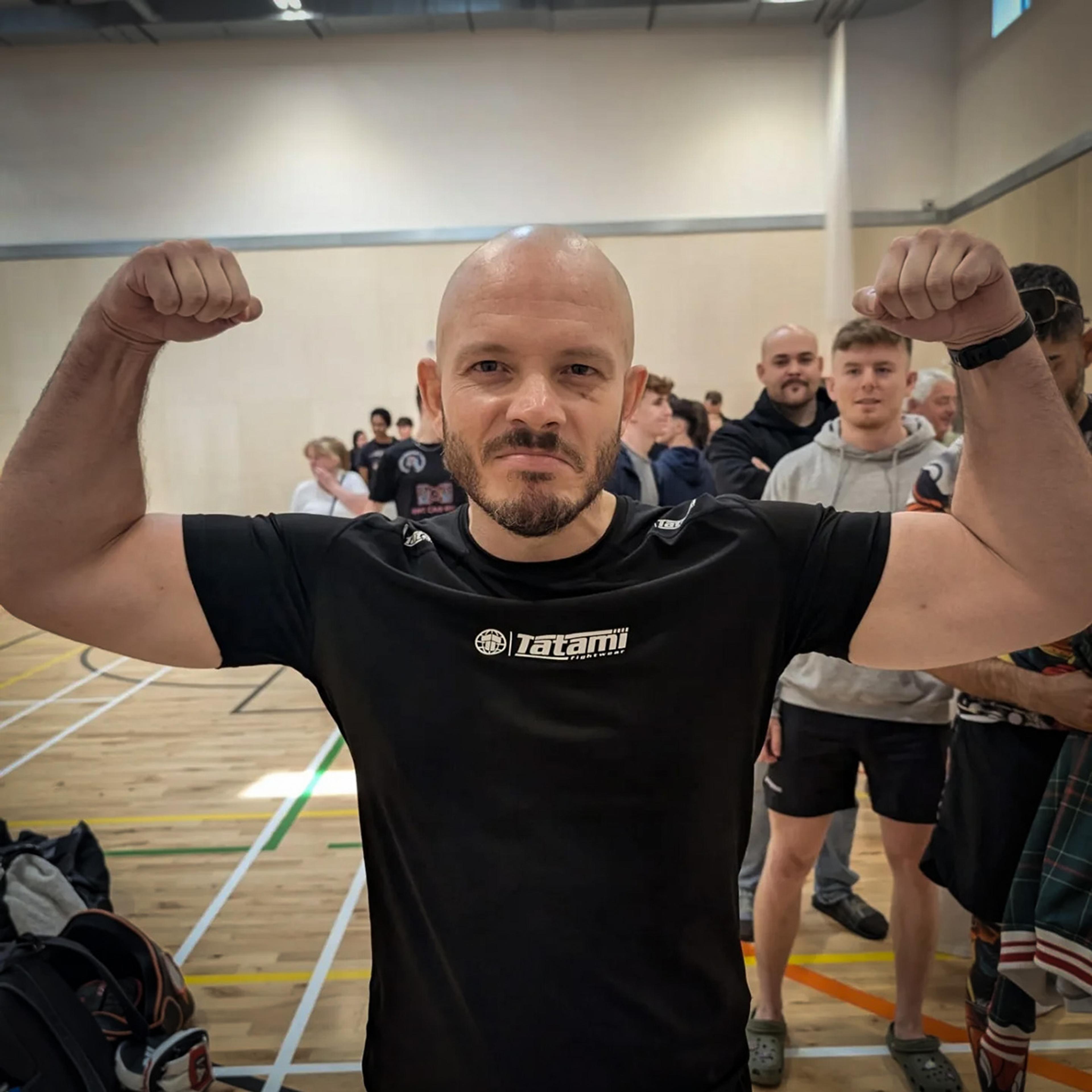 A man in a gym flexing his arms towards the camera with people in the background on a sports court.