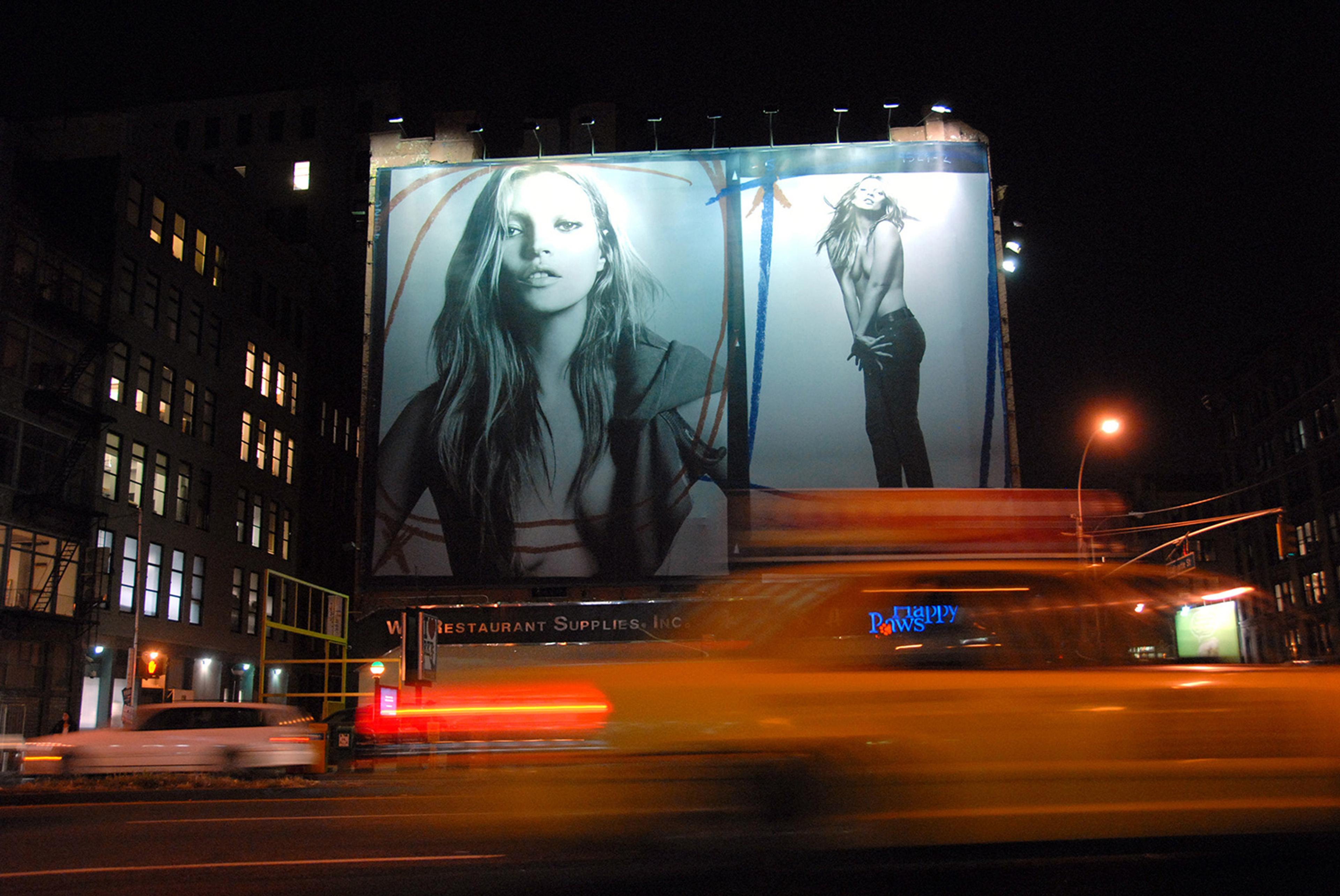 Night photo of a city street featuring a large billboard with a black and white fashion advertisement and blurred taxi lights.