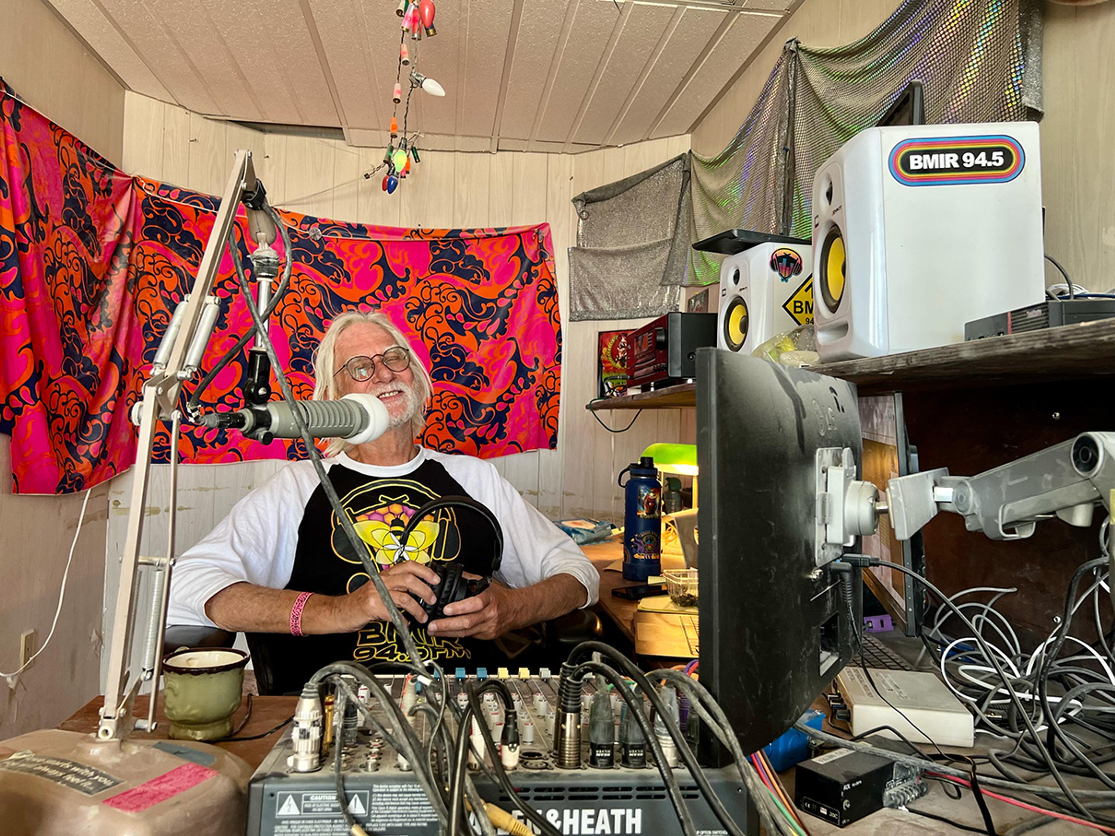 A smiling man with headphones in a colourful radio studio, surrounded by microphones and audio equipment.