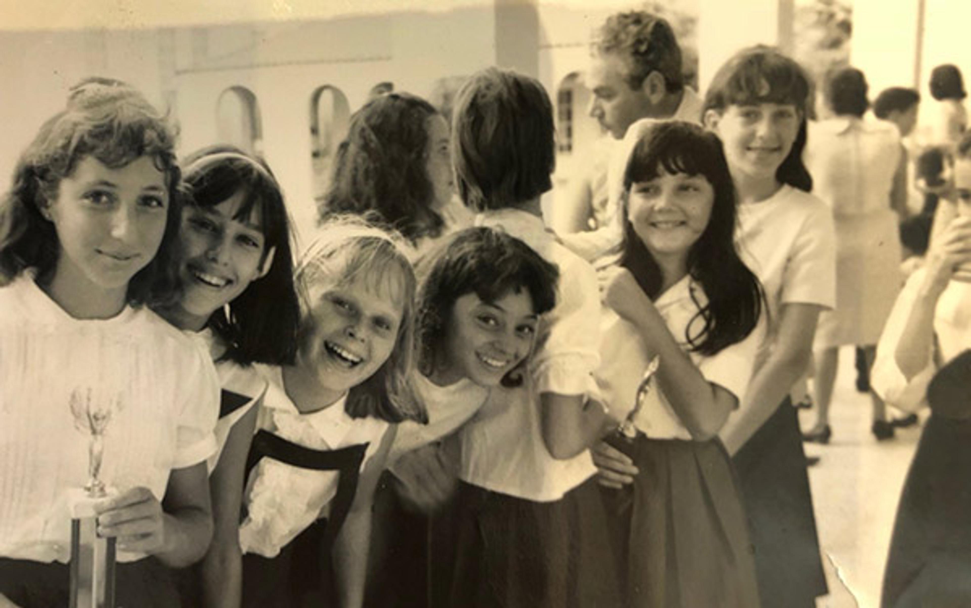 Vintage photo of young girls in school uniforms smiling and standing in line, with adults in the background.