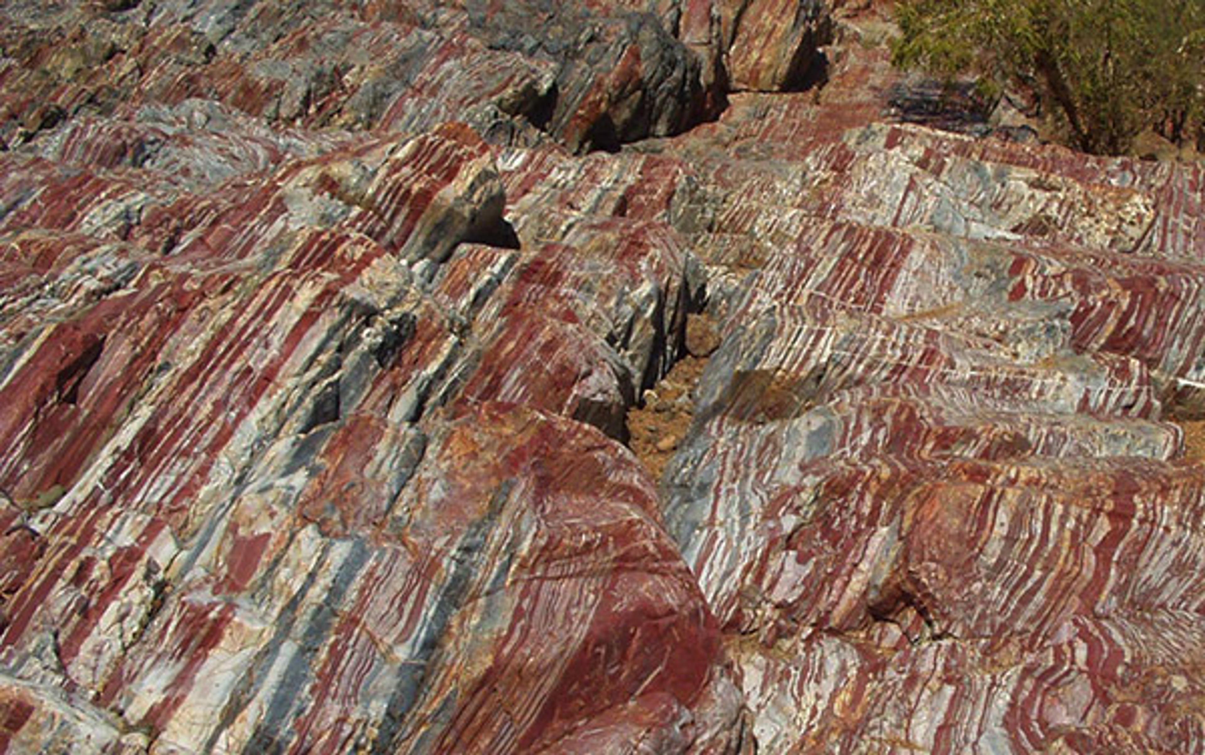 Colourful layered rock formations with red, brown and white stripes. Sparse vegetation is visible in the background.