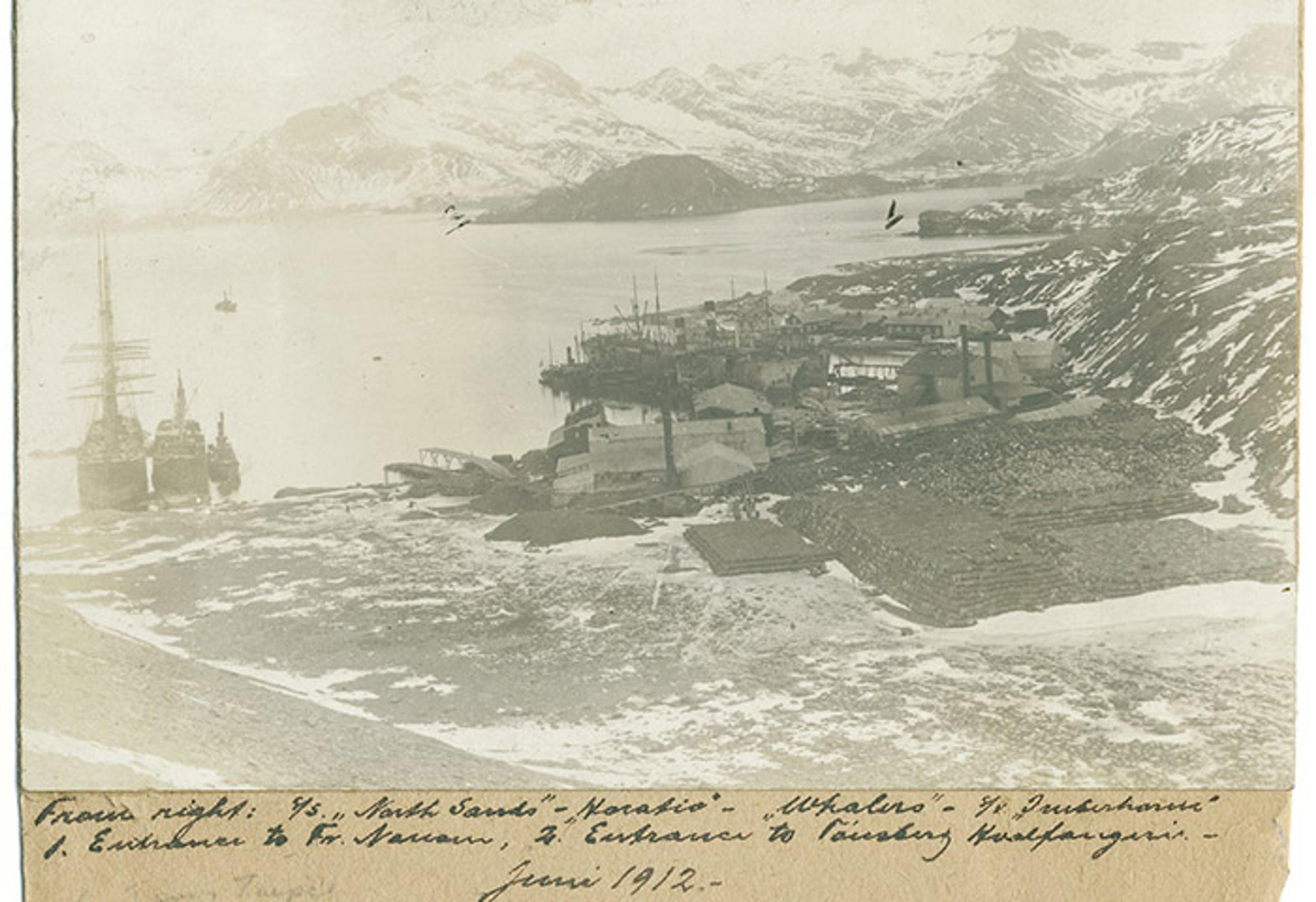 A sepia photo of a coastal settlement with ships and snowy mountains in the background, captioned “June 1912” at the bottom.