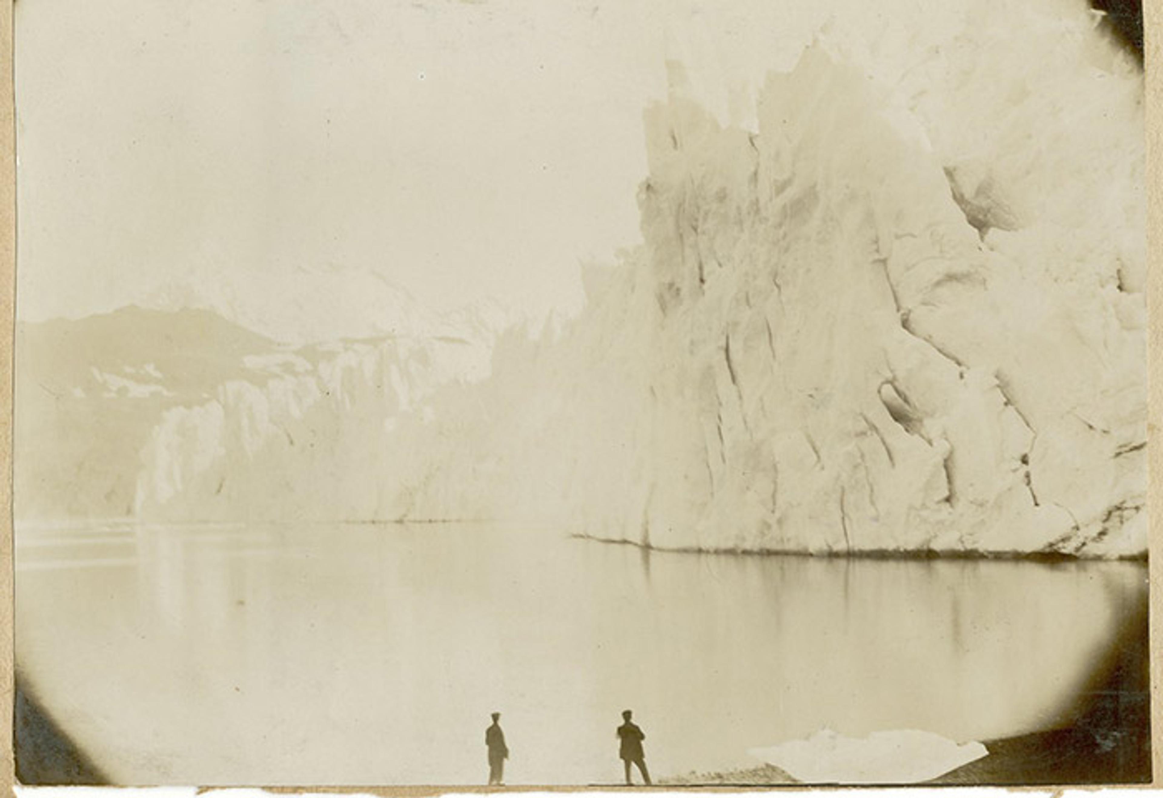 Vintage photo of two silhouetted figures standing by a lake with large ice cliffs in the background.
