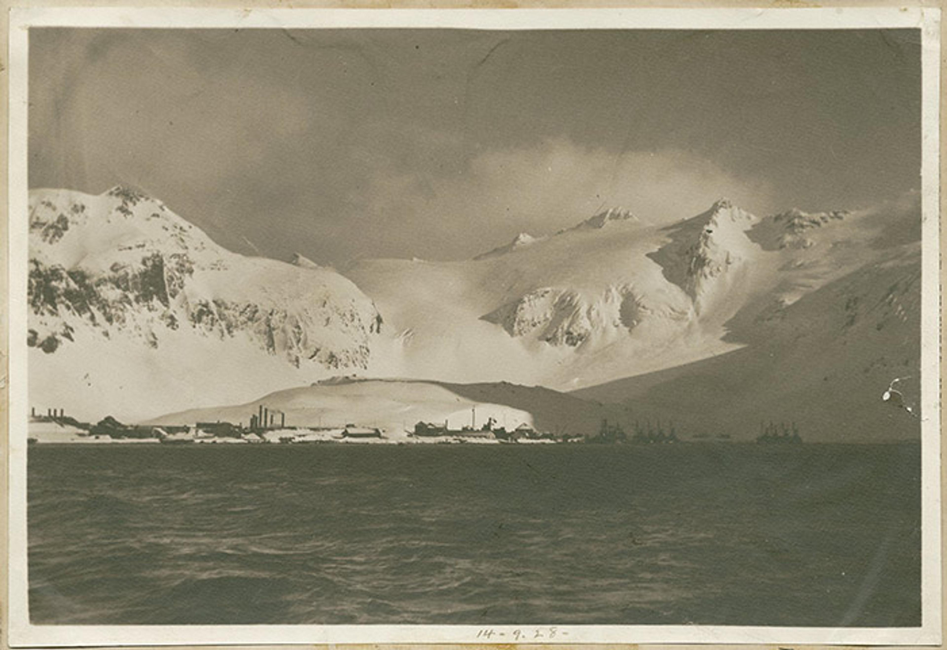 Vintage photo of a snow-covered industrial settlement at the base of rugged mountains by a body of water, dated 14-9-38.