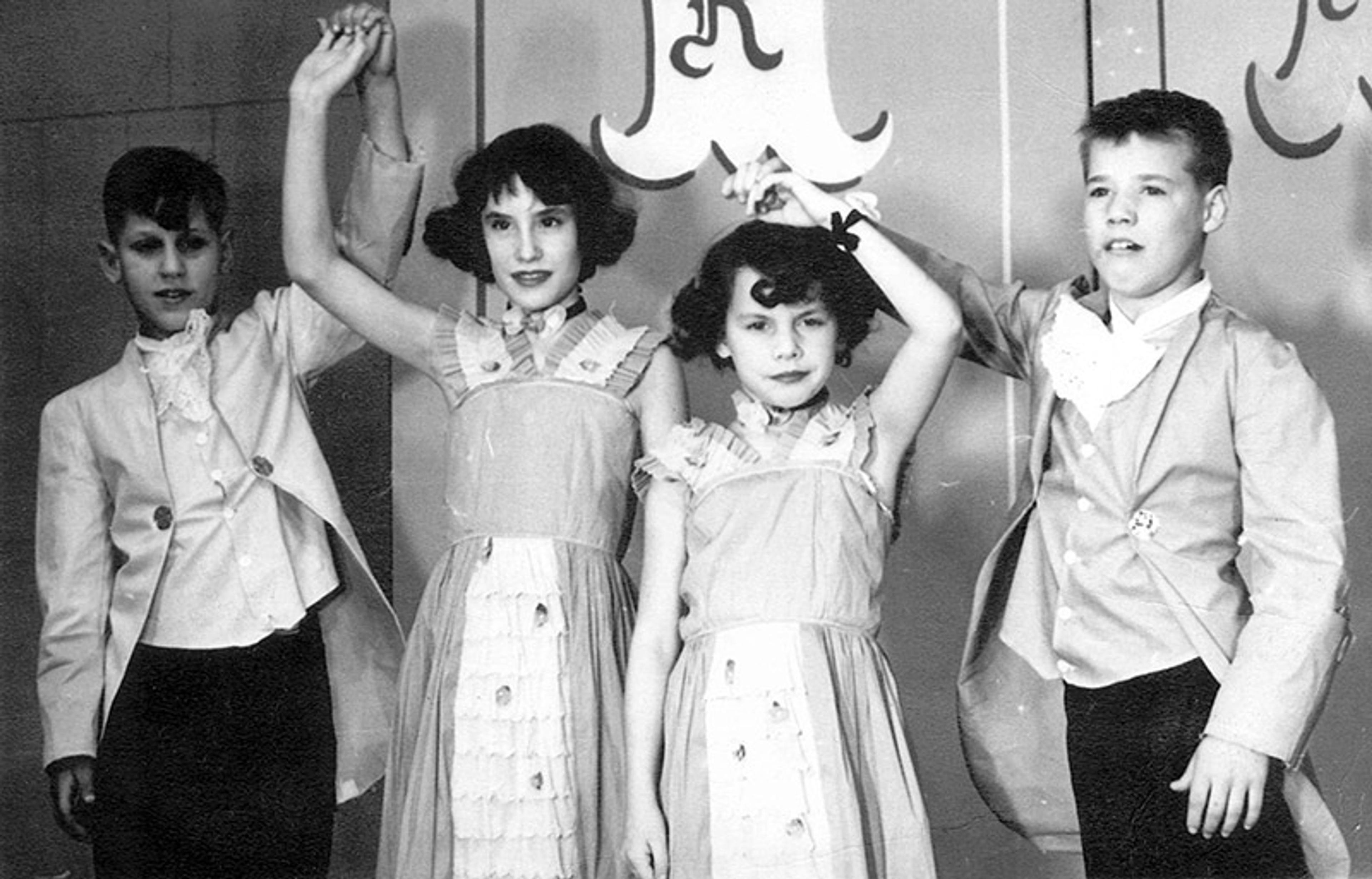 Vintage photo of two boys and two girls in formal attire holding hands in pairs, standing in front of a decorated wall.