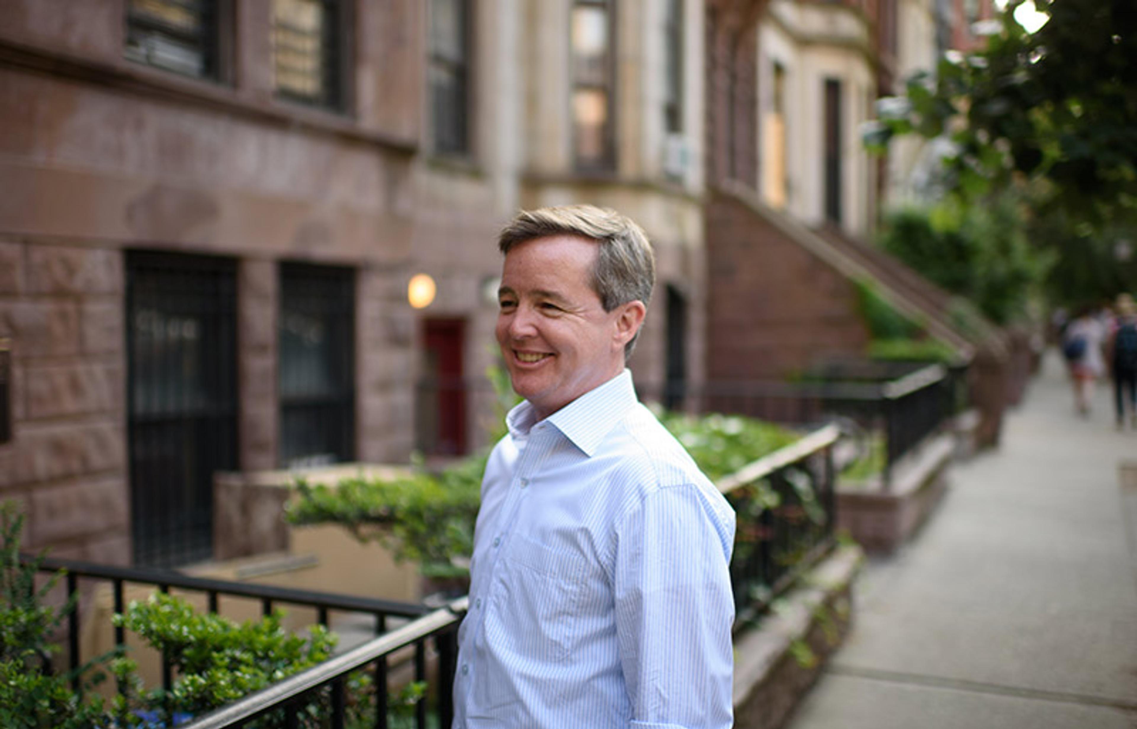 A smiling man in a white shirt standing on a tree-lined street with brownstone buildings in the background.