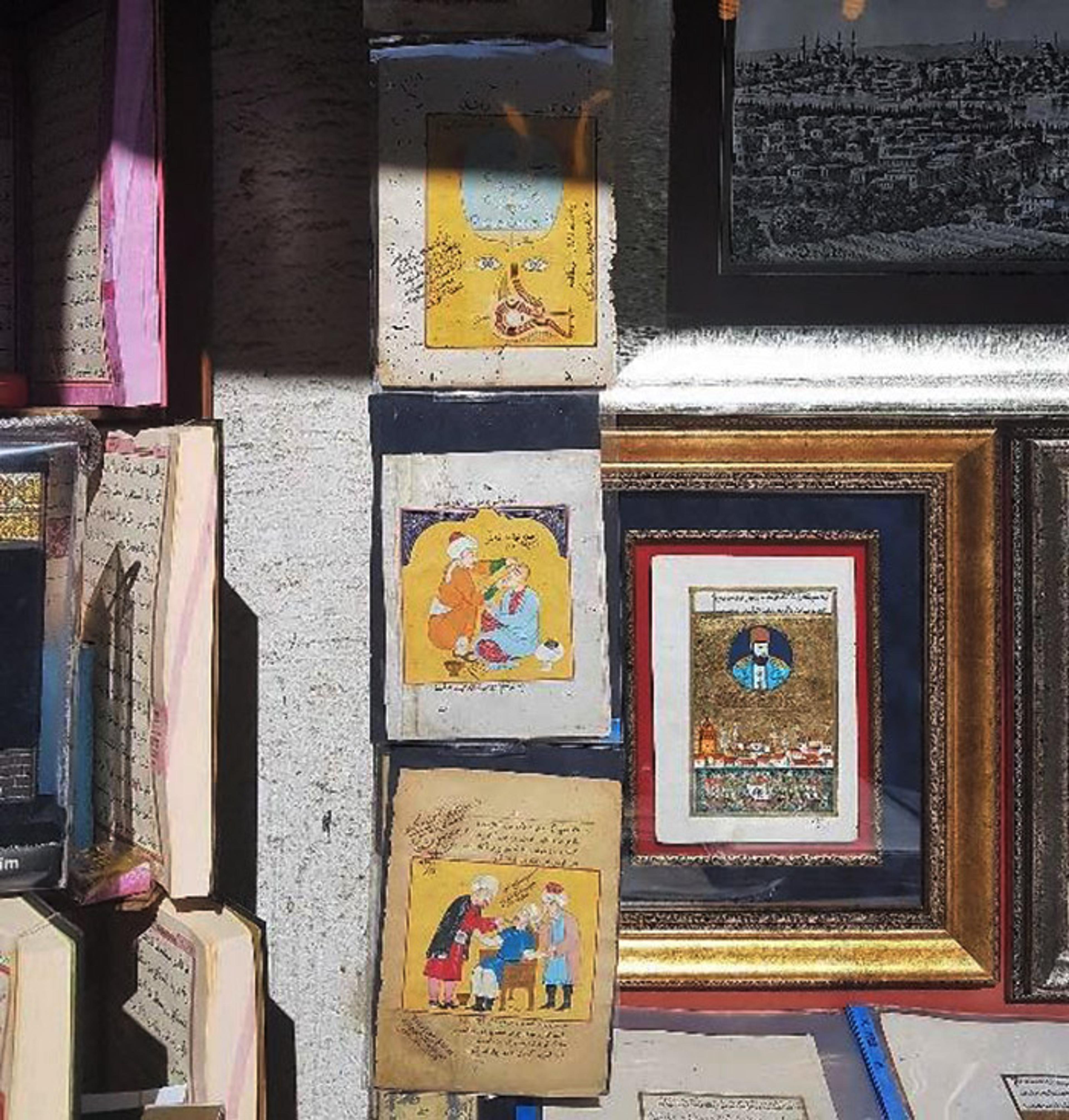 Framed artworks and pages with illustrations and text, likely old manuscripts, displayed on a wall with other books.
