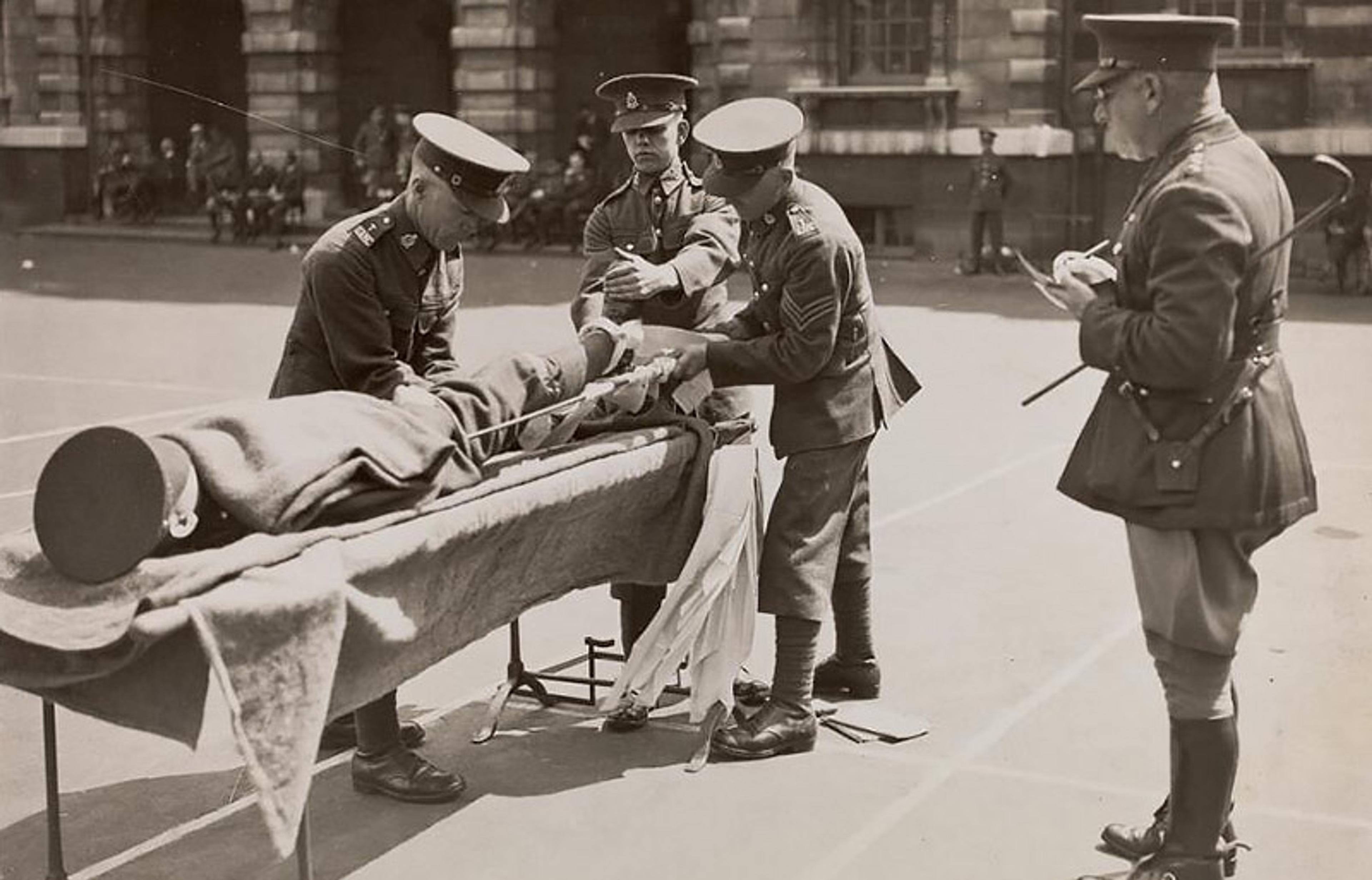 Black and white photo of soldiers practising medical procedures on a dummy on a stretcher outdoors with an officer observing.