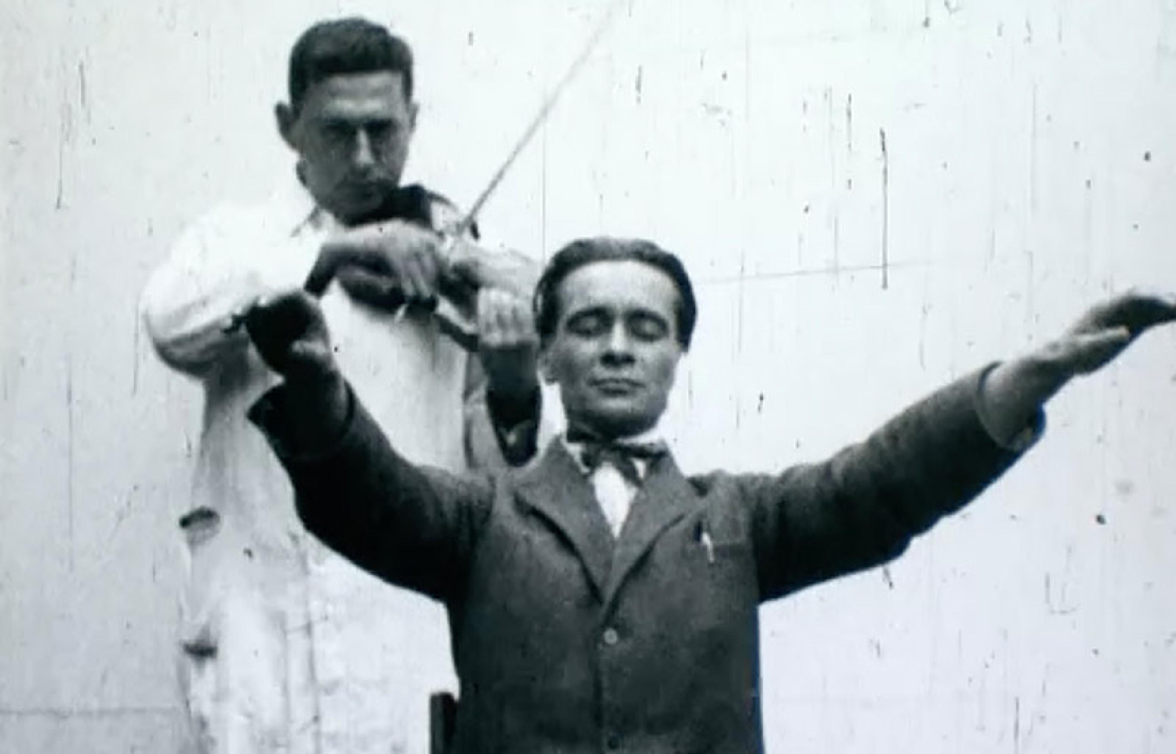 A vintage black and white photo of a man in a lab coat playing the violin behind another man whose arms are stretched in front of him, eyes closed.