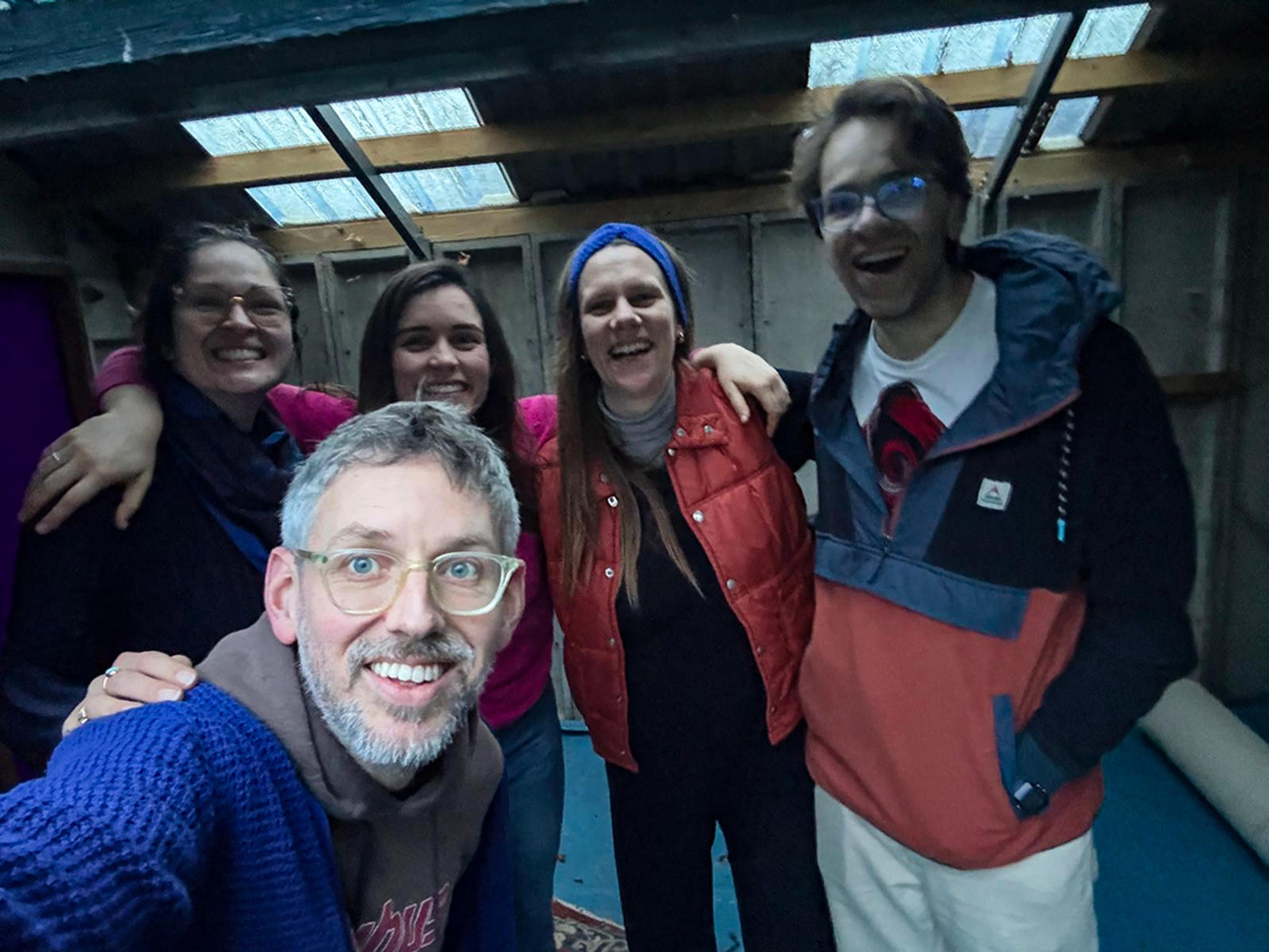 Five smiling people wearing casual clothing in a dimly lit room with skylights. One person is taking a selfie.
