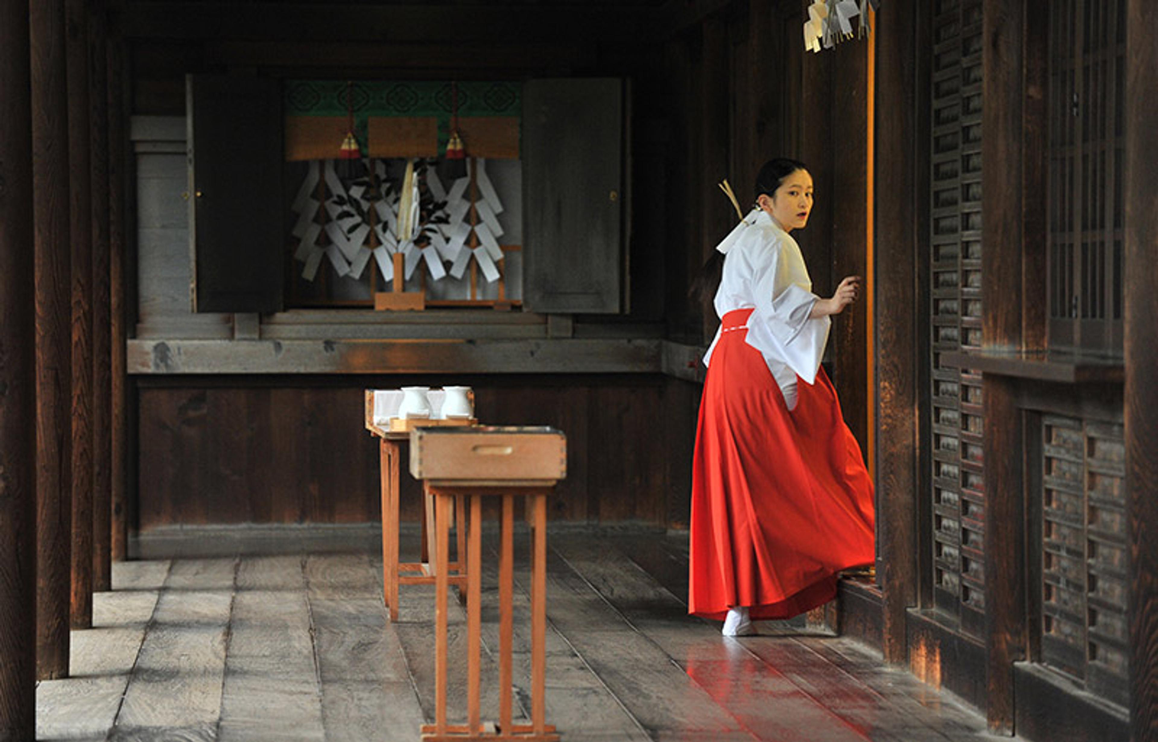 A woman in traditional Japanese attire in a wooden shrine with white and red decor.
