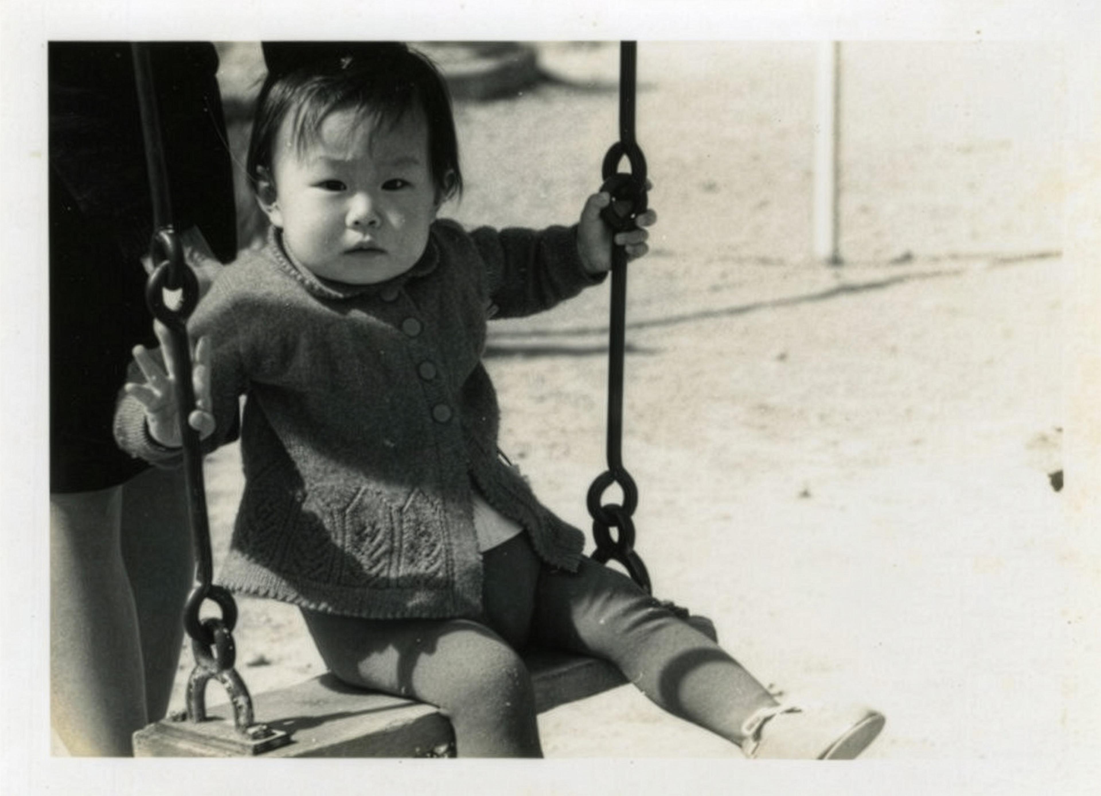 Black and white photo of a toddler on a swing wearing a knitted cardigan with an adult standing nearby.